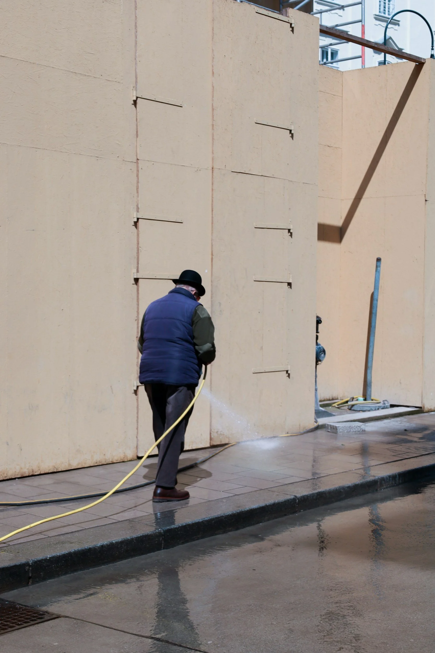 A person wearing a black hat, blue vest, and green sleeves is pressure washing a sidewalk in front of a large beige wall. Water and soap are visible on the ground, and the person is using a yellow hose connected to a pressure washer.