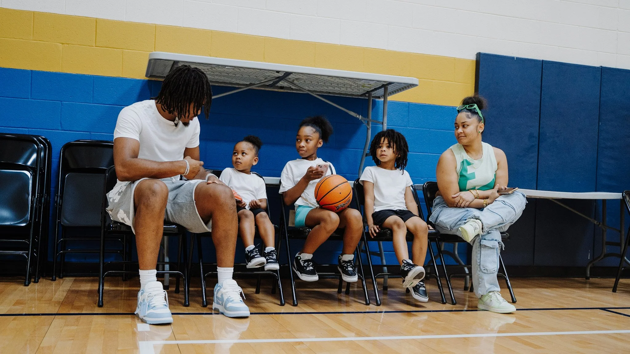 A group of children and two adults sitting on chairs in a gymnasium with blue and yellow walls. The children are holding a basketball and a phone, and the adults are sitting with them. One adult is a man with dreadlocks, and the other is a woman with sunglasses on her head.