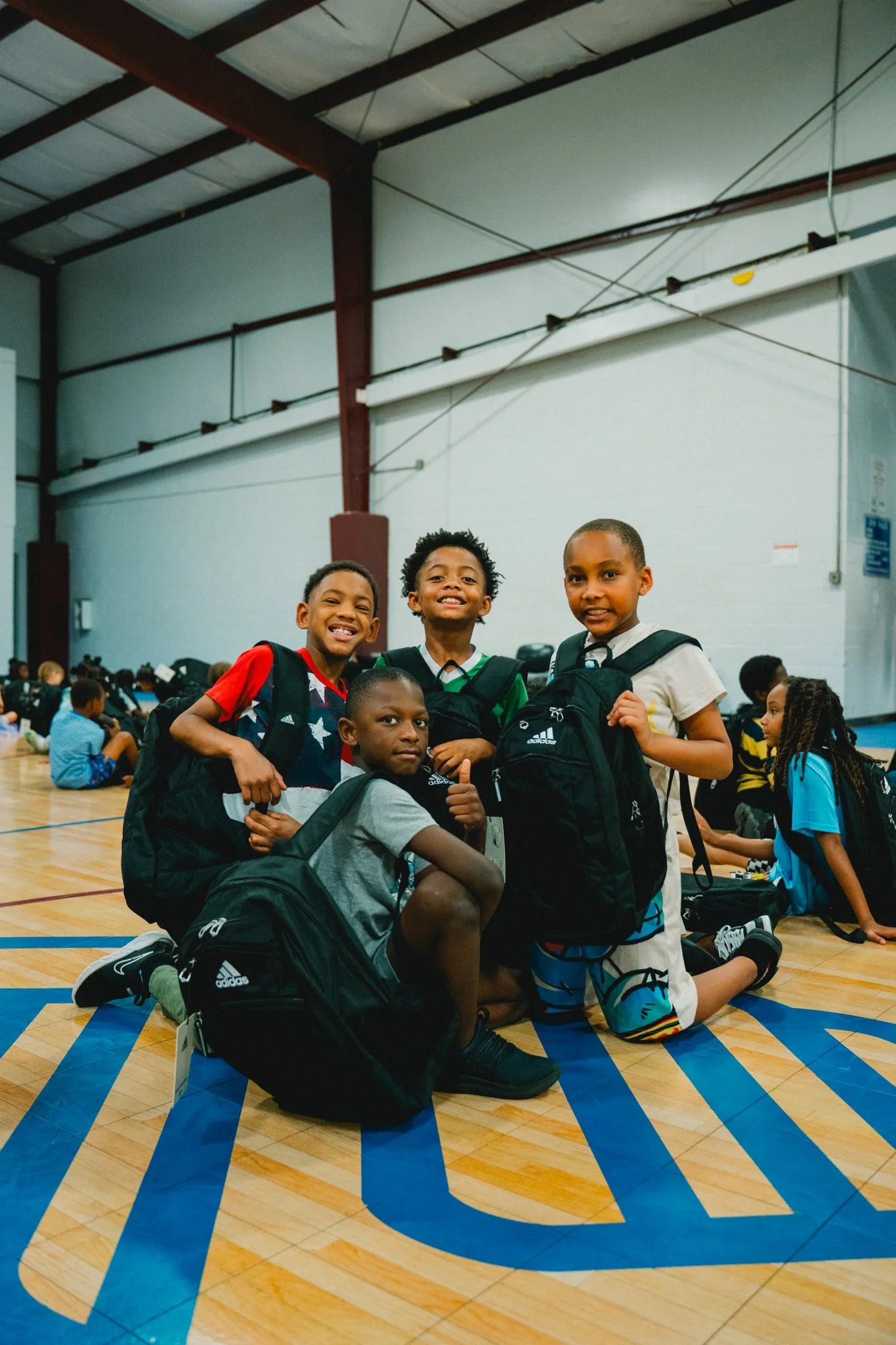 A group of boys with backpacks kneeling and standing on a basketball court, smiling at the camera inside a gymnasium filled with other children.