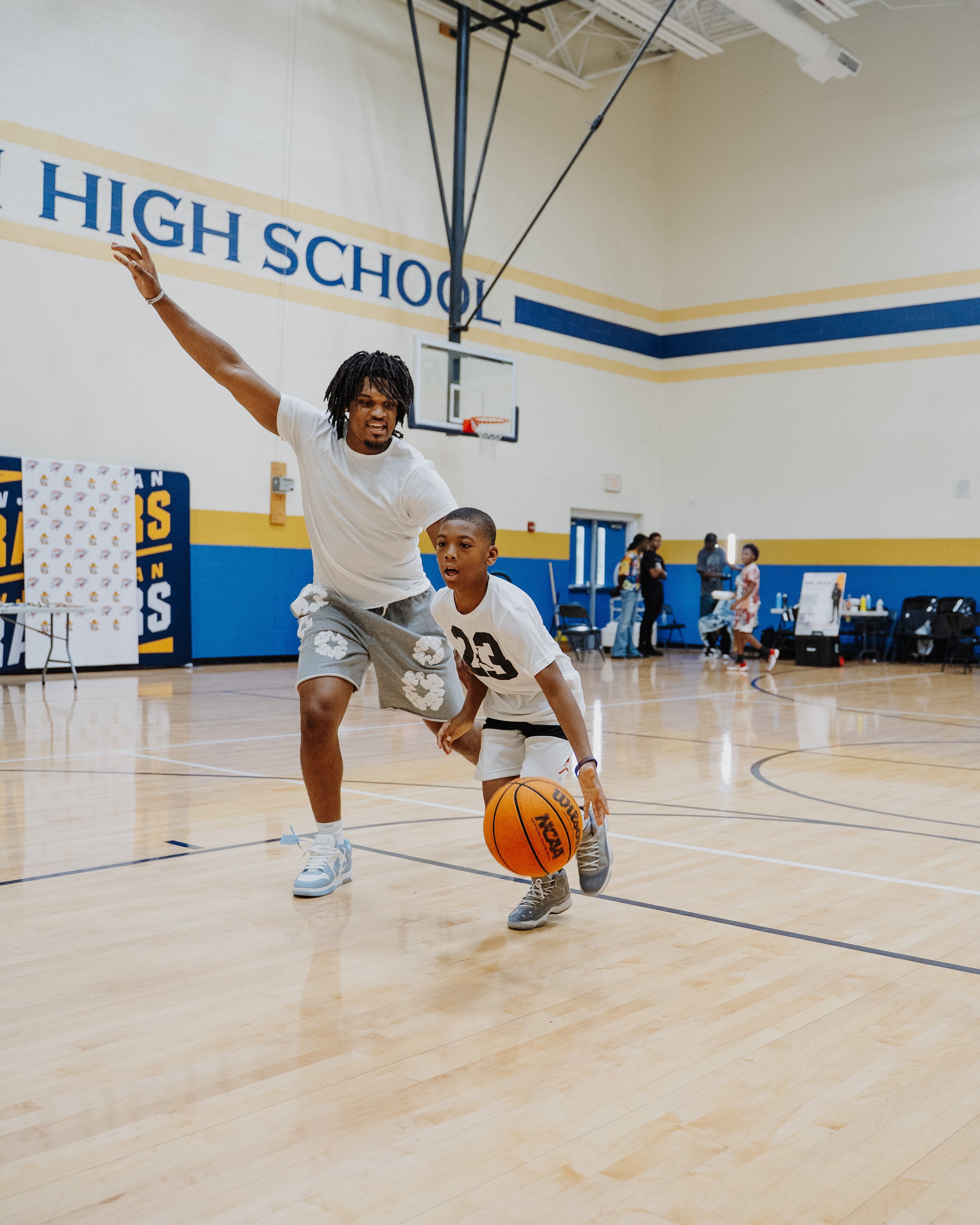 A man and a boy playing basketball in a gymnasium. The man appears to be defending the boy as he dribbles a basketball. The gym has a basketball hoop and high school lettering on the wall.
