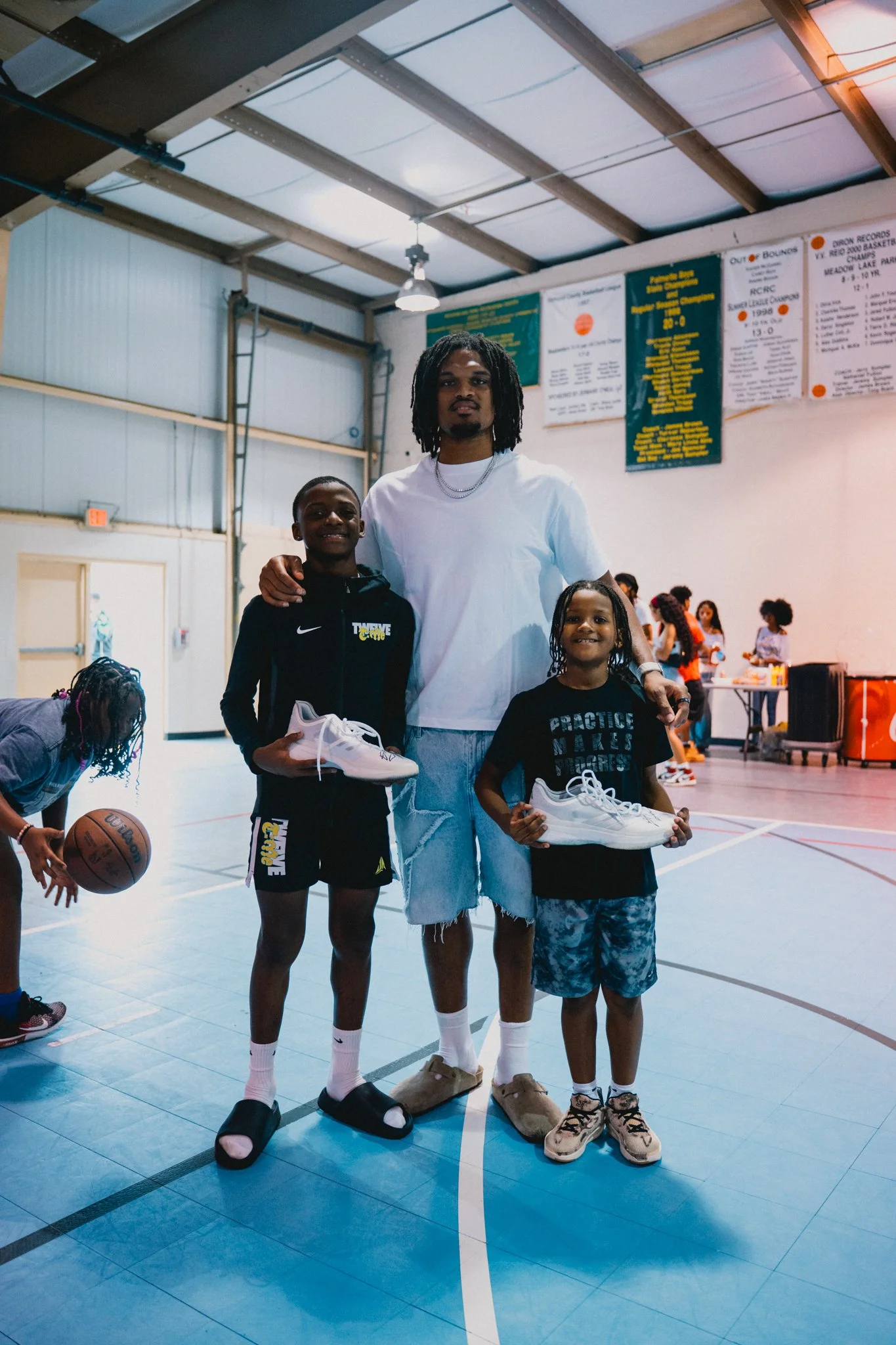 A man and two children standing in an indoor gymnasium, holding shoes. The man has dreadlocks and wears a white t-shirt and ripped jeans. The girl wears a black hoodie, shorts, socks, and slippers, while the boy wears a black t-shirt, shorts, socks, and sandals. There are other children in the background, and banners hanging on the wall.