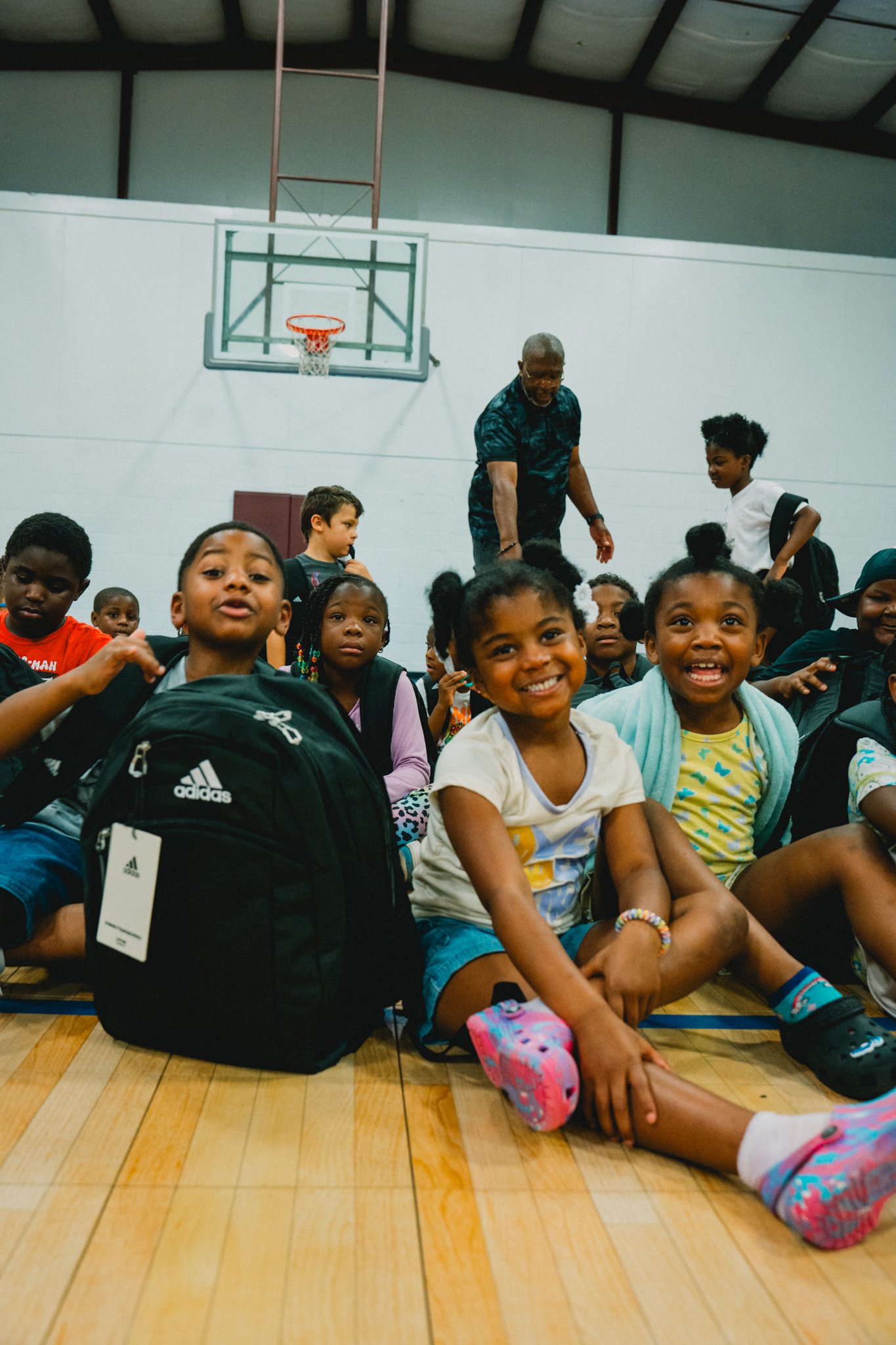 A group of children sitting on the wooden gym floor, smiling and looking at the camera, with an older man standing behind them in a gymnasium with a basketball hoop and high ceiling.