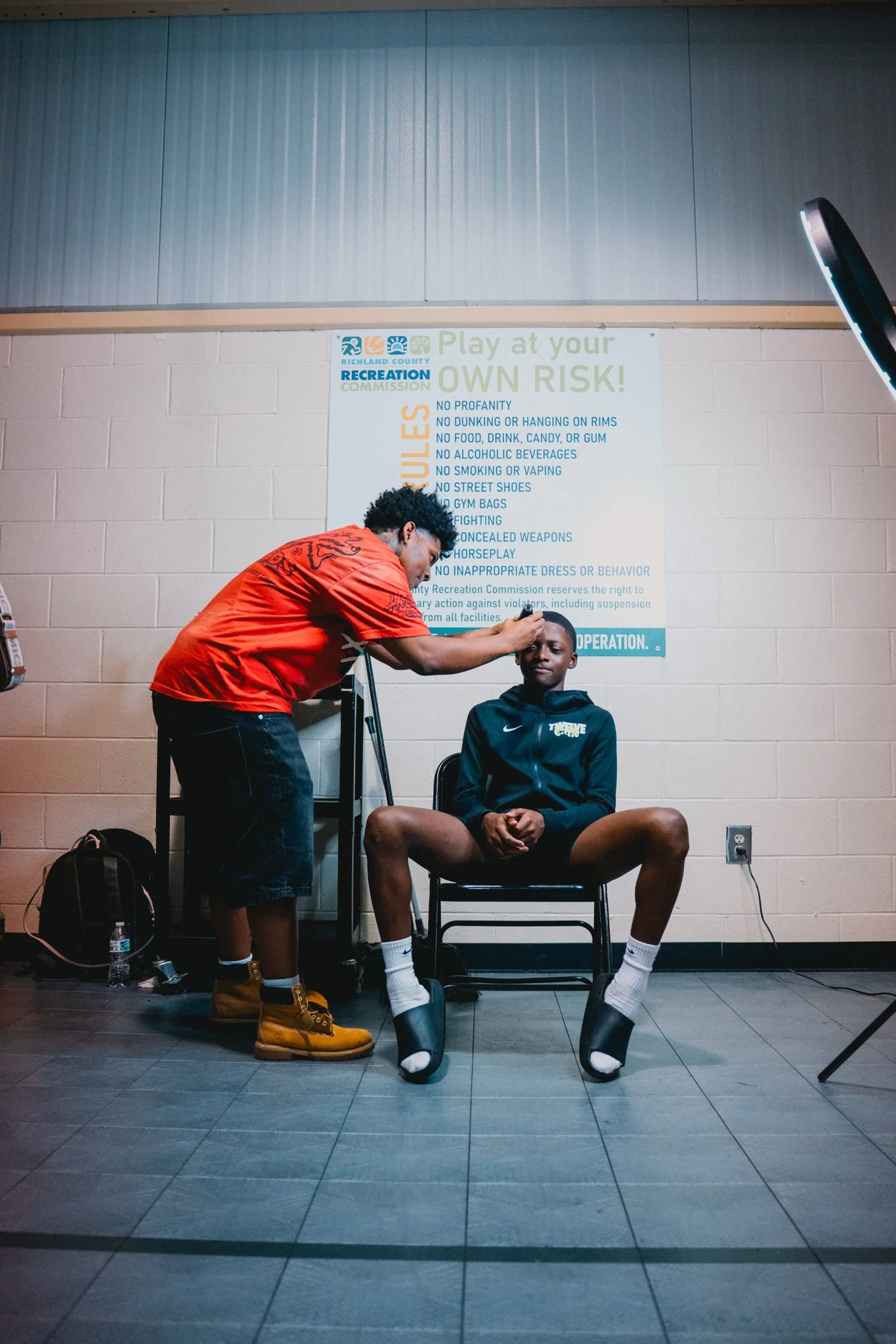 A person with curly hair and a red jacket is applying makeup or checking on a young man sitting on a chair with his legs apart, wearing a black Nike jacket and black shorts, in a room with a beige cinder block wall and a large sign behind them. The sign lists rules for a recreation center.