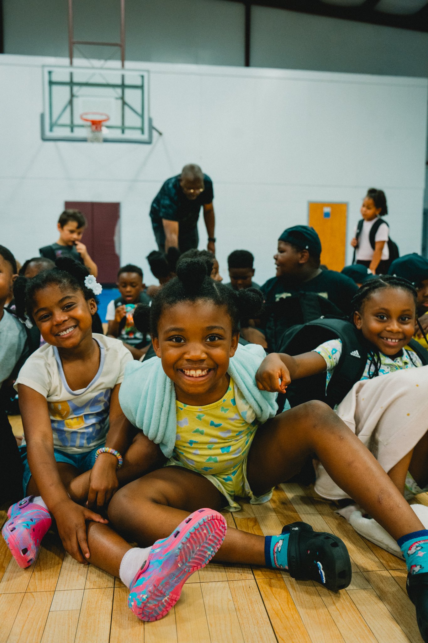 A group of smiling children, mostly girls, sitting on a gymnasium floor. In the background, a basketball hoop and gym wall are visible.