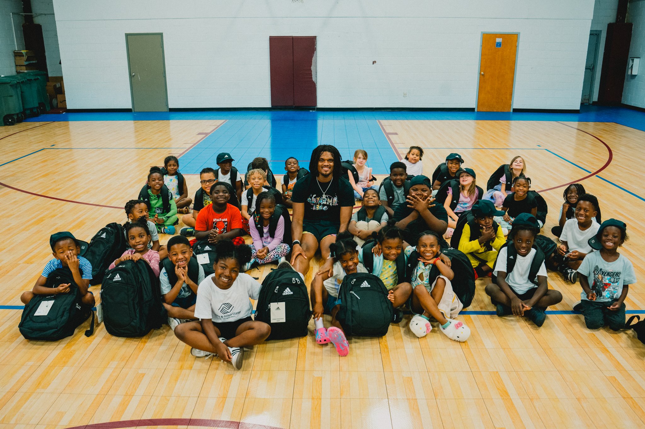 A large group of children sitting on the gymnasium floor with a man in the center, all smiling and posing for the photo. The children are wearing backpacks and casual clothing, and the gym has a wooden floor with blue lines.