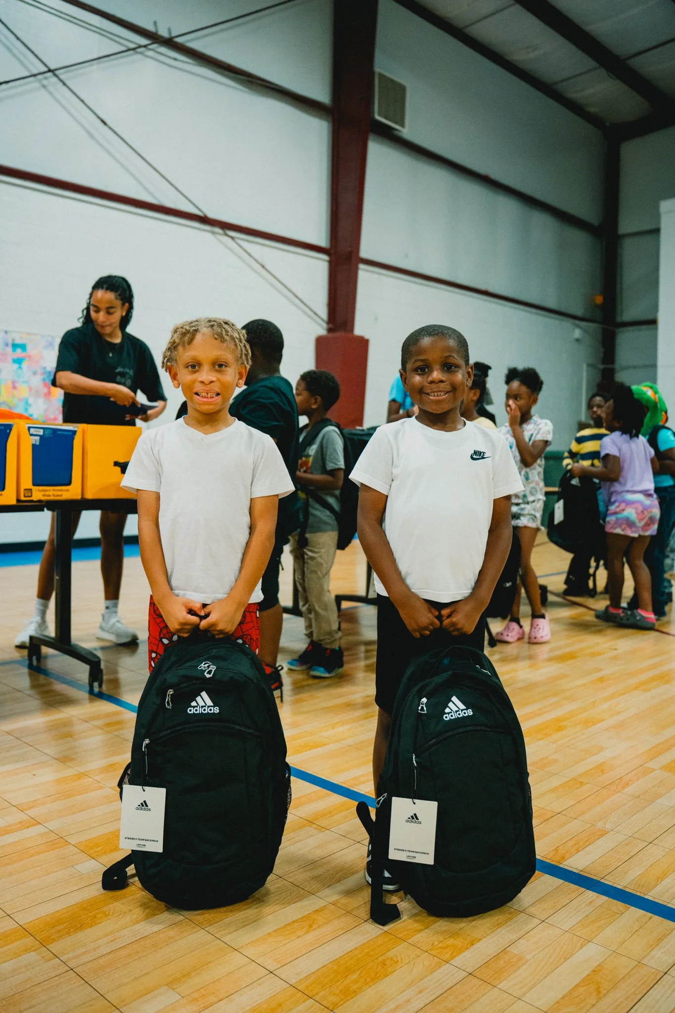 Two young boys with backpacks standing in a gymnasium, smiling at the camera, with children and a woman in the background.