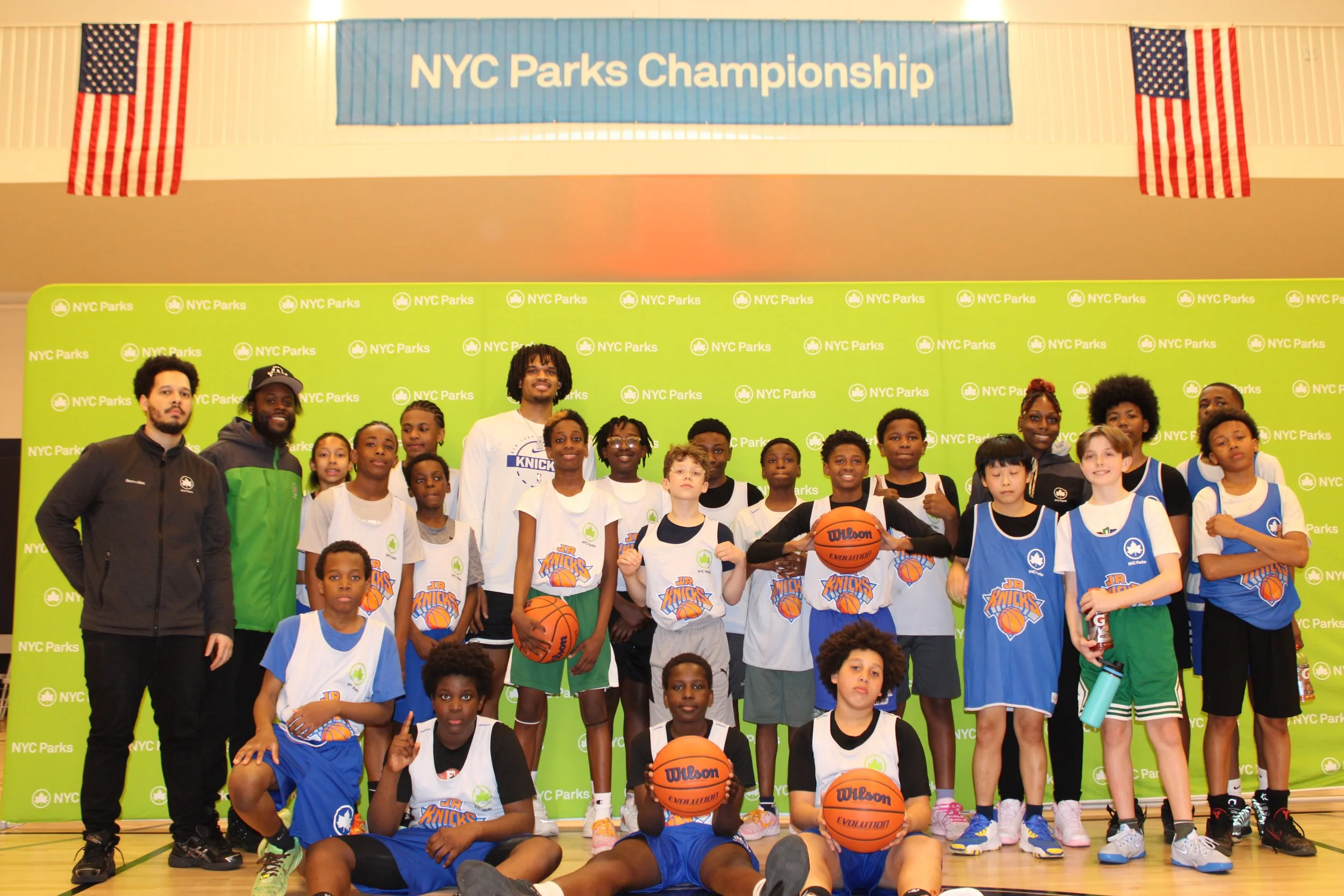 Children and coaches in a group photo during a NYC Parks basketball event, with a banner reading 'NYC Parks Championship' and American flags in the background.