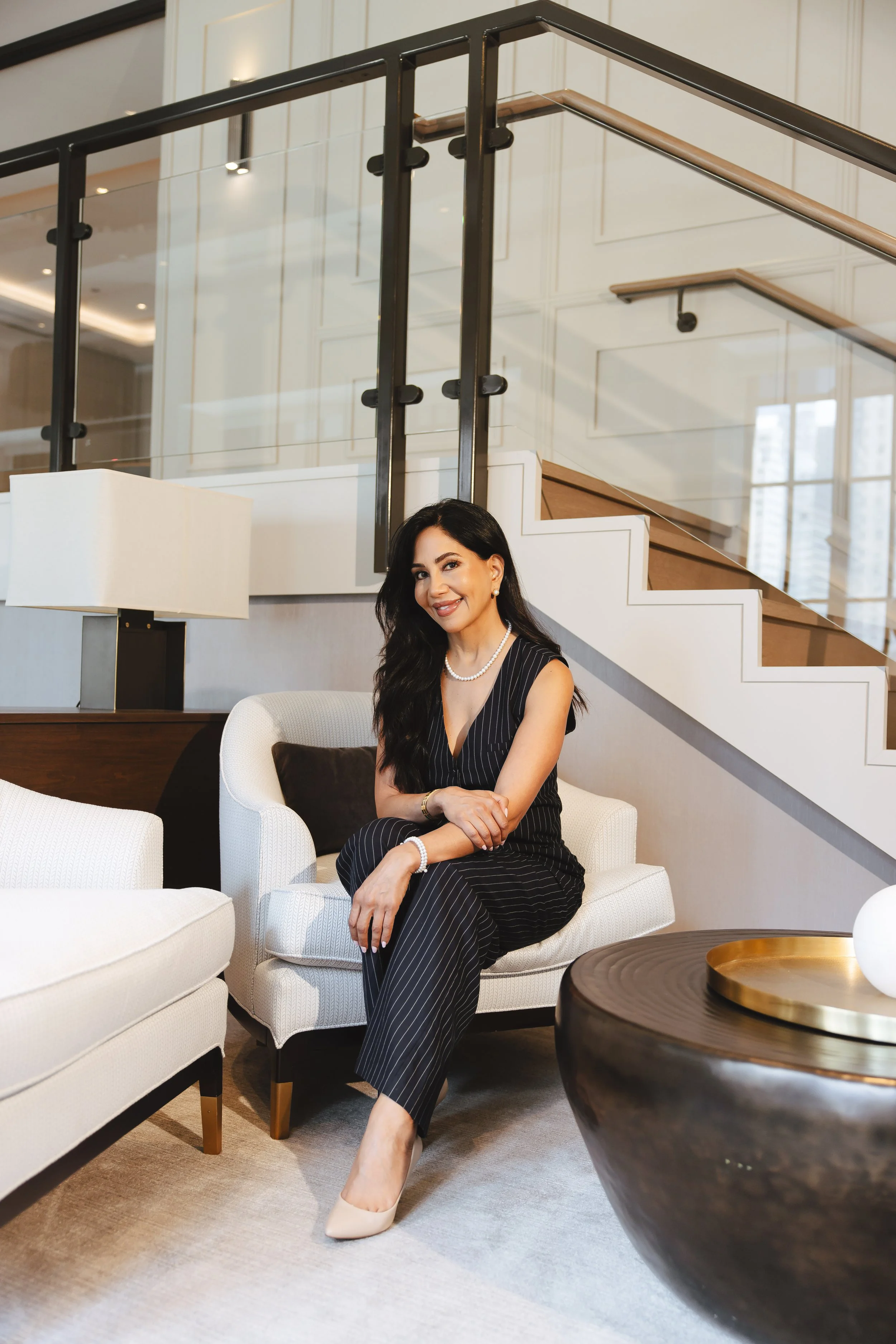 A woman in a black pinstripe jumpsuit with pearl jewelry sitting on a cream armchair in a modern living room with white walls, glass staircase, and a black and gold side table.