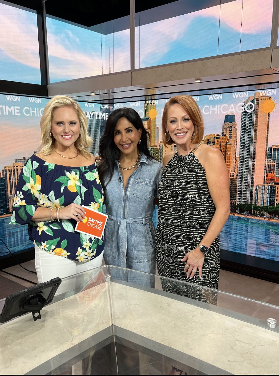 Three women standing in a TV studio with a Chicago skyline backdrop, smiling at the camera. One woman is holding a card that says 'Daytime Chicago.'