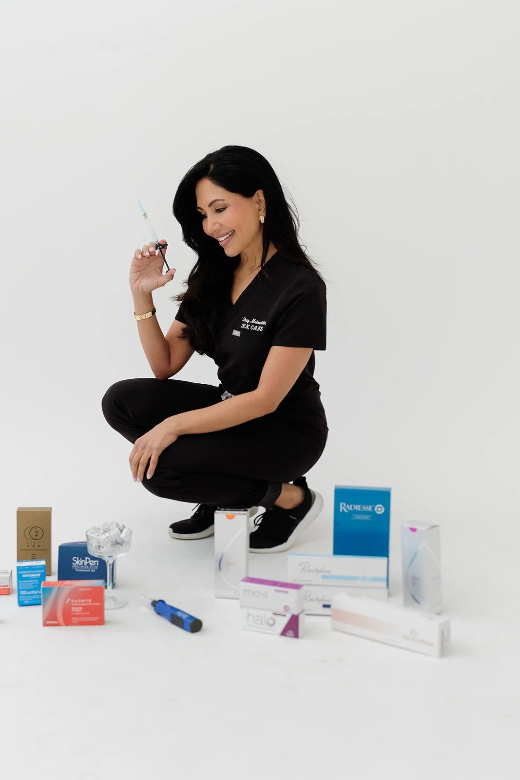 A woman in black scrubs crouches on the floor, holding a syringe and smiling. Various healthcare products are arranged around her on the floor.