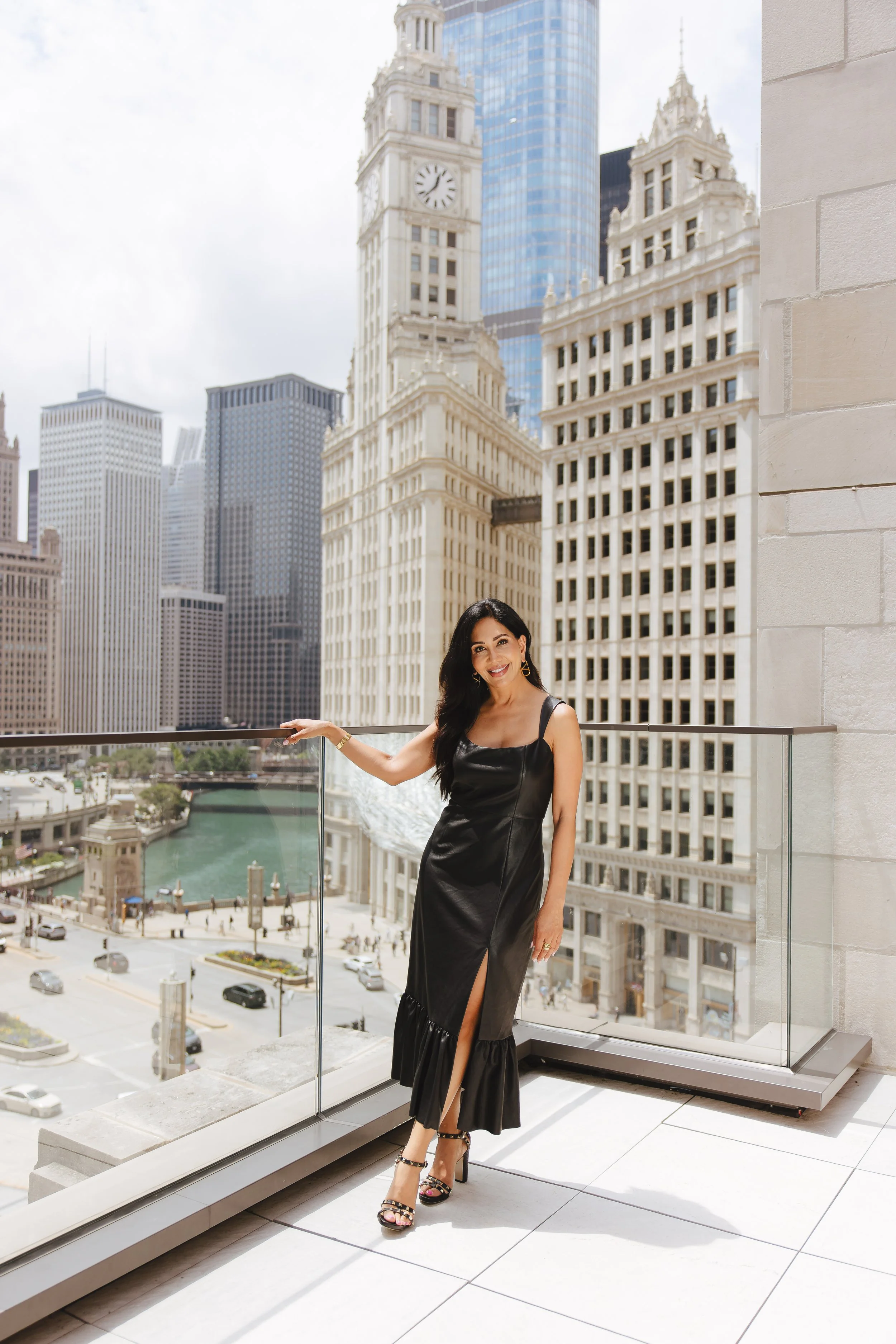 A woman in a black dress with a high slit stands on a balcony overlooking a cityscape with tall buildings, a river, and a busy street.