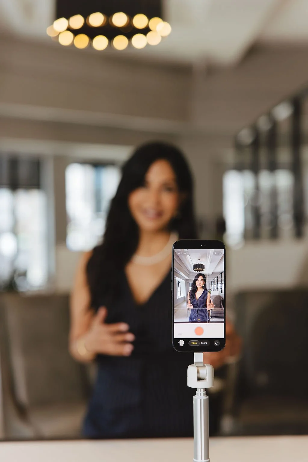 A woman is smiling while taking a selfie with a smartphone on a selfie stick in a well-lit indoor space with large windows and modern decor.