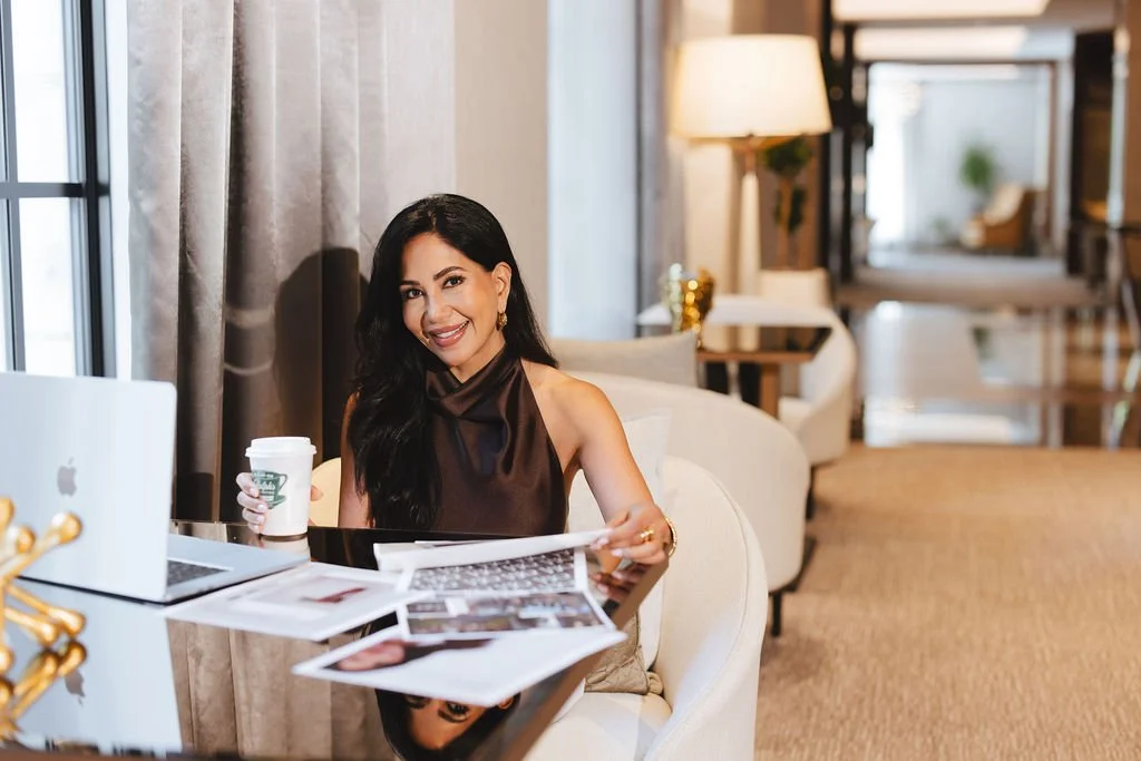 A woman with long dark hair, wearing a sleeveless brown top, sitting at a desk with a laptop, coffee cup, and papers, smiling in a well-lit modern office or lounge.