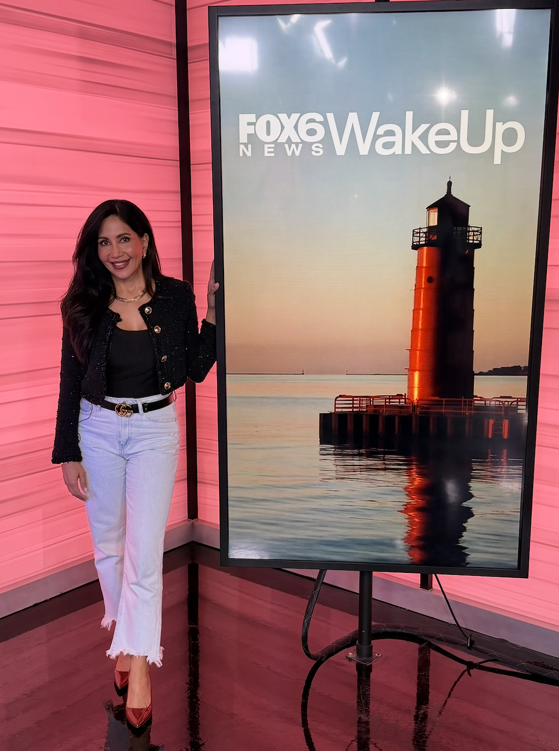 Woman standing next to a large television screen displaying a lighthouse at sunset, with a pink wall in the background.