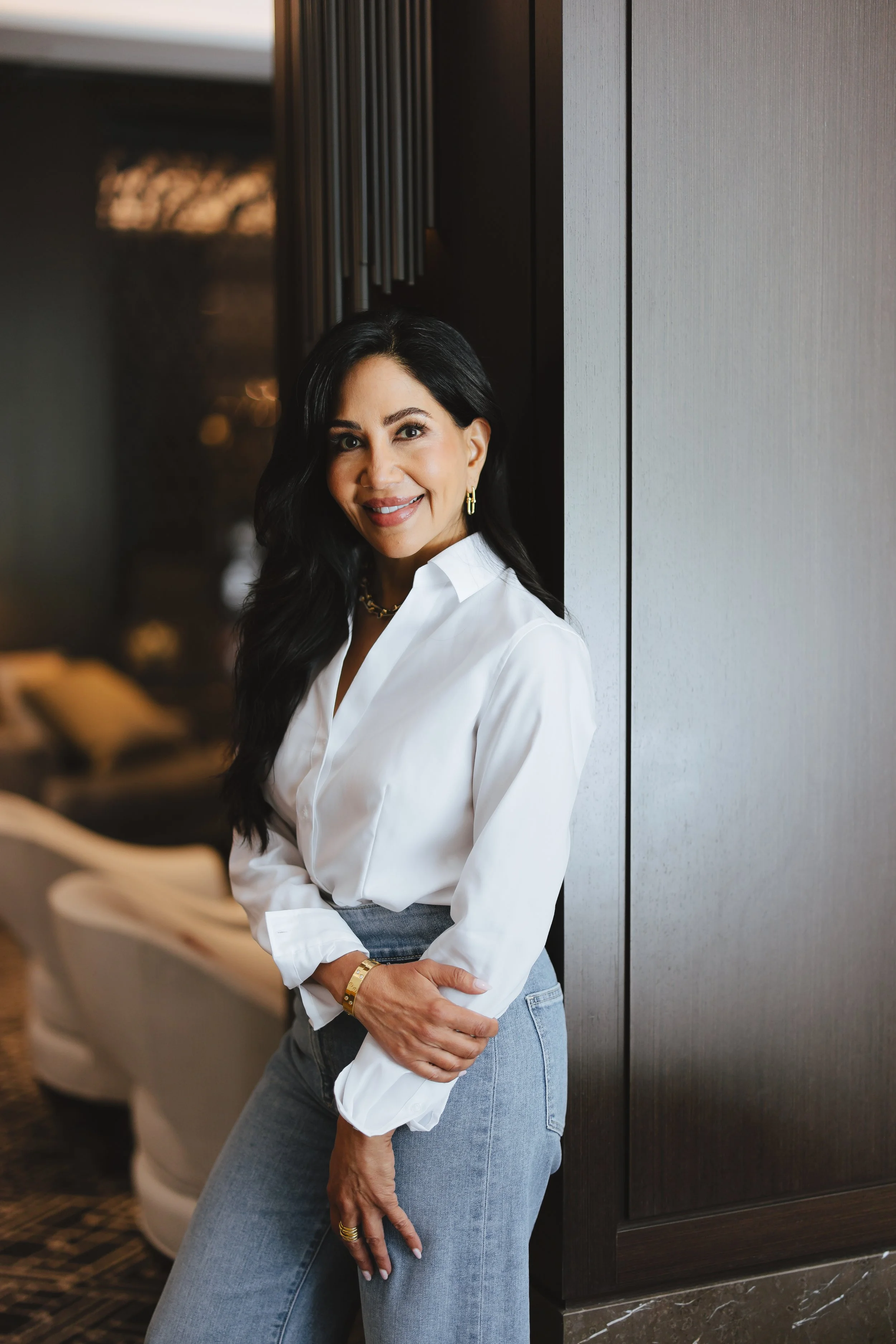 A woman with long black hair, wearing a white shirt and light blue jeans, is leaning against a dark-colored wall in a modern interior space, smiling at the camera.