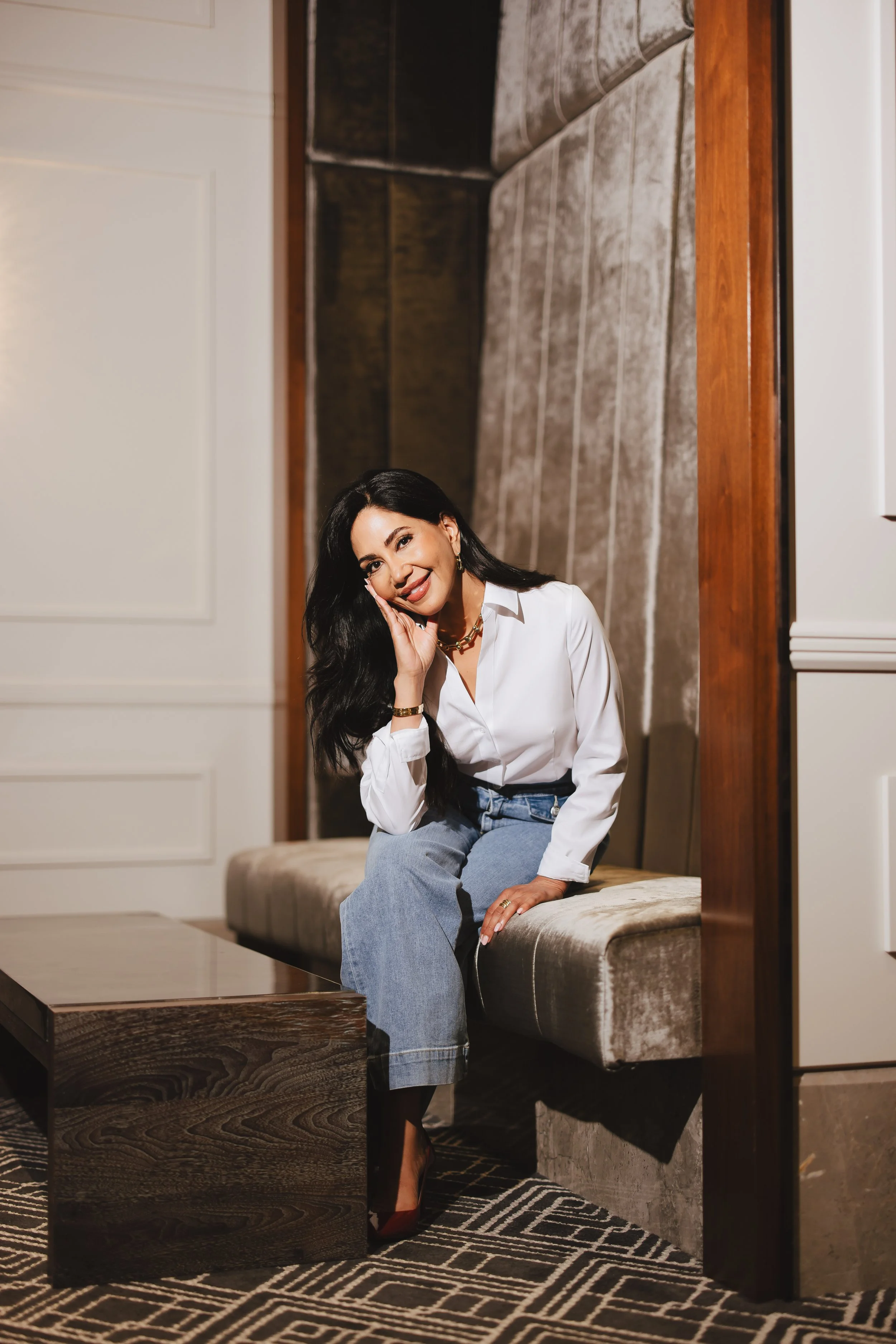 A woman with long dark hair, wearing a white blouse and light blue jeans, is sitting on a beige velvet bench in a stylish interior space. She is smiling and resting her head on her hand, looking at the camera.