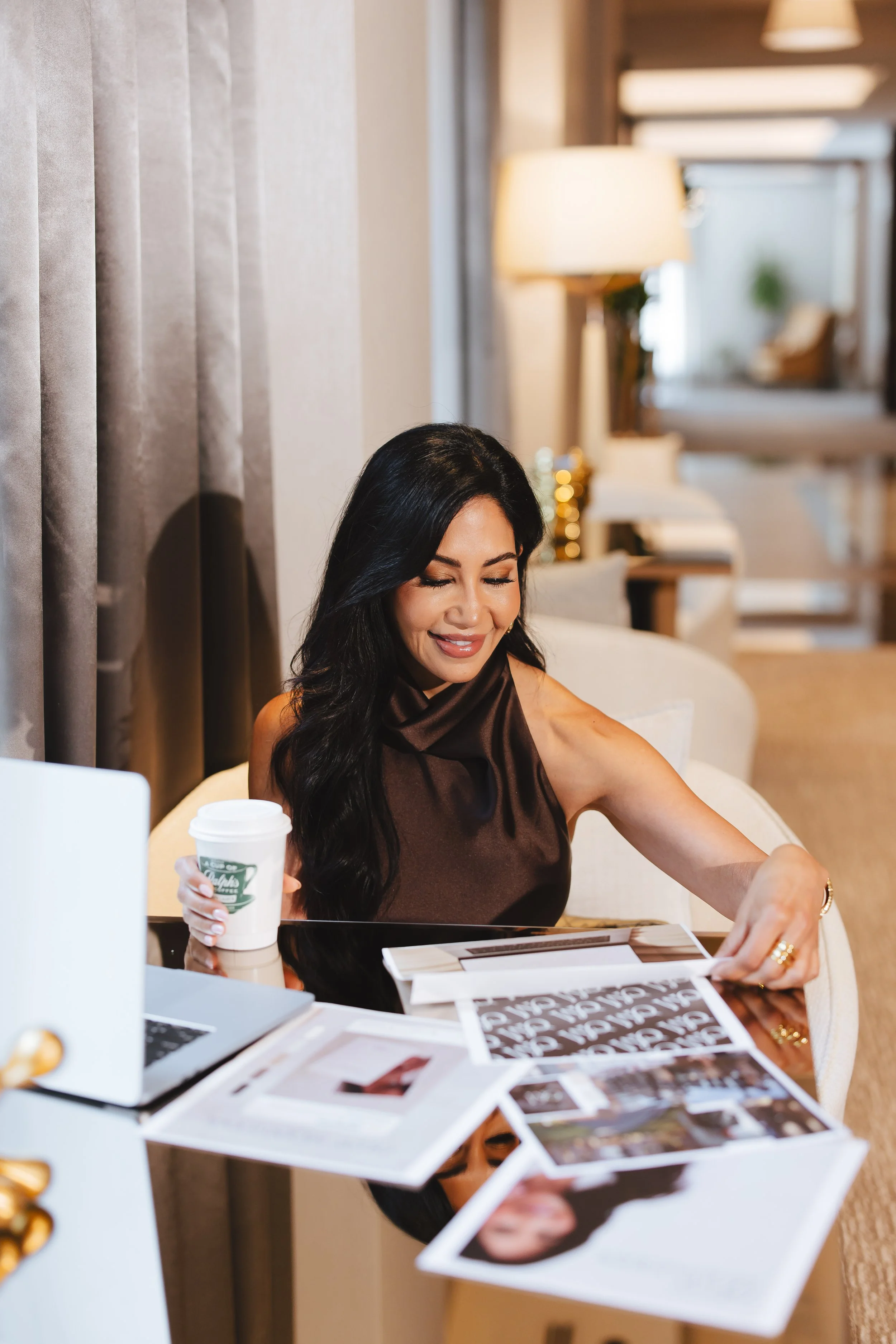 A woman with long dark hair, wearing a sleeveless brown top, is sitting at a table and smiling while looking at magazines or catalogs. She is holding a white paper cup with a green logo. The background shows a cozy, well-lit room with lamps, curtains, and decorative elements.