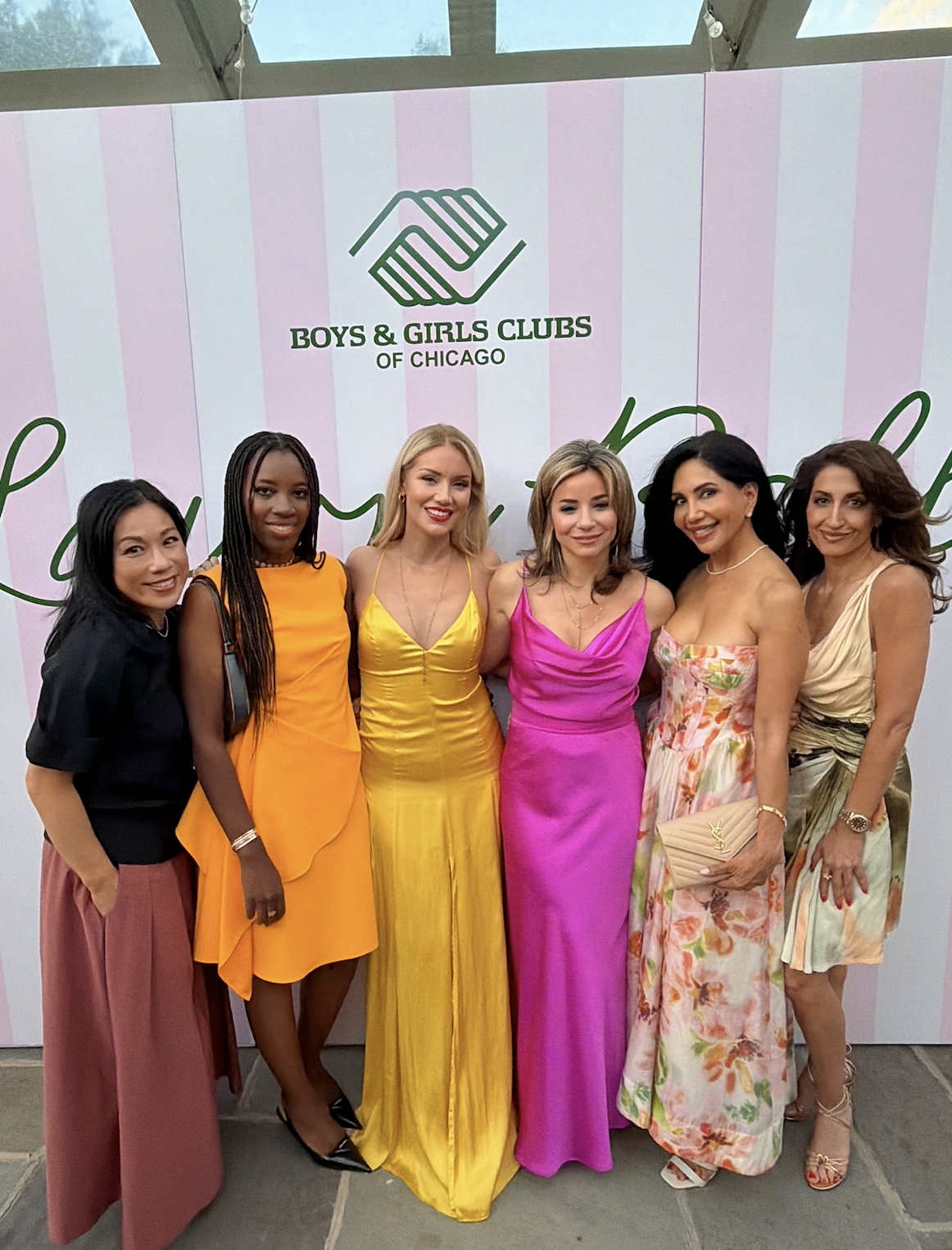 Group of six women standing together at a Boys & Girls Clubs of Chicago event, posing for a photo in front of a pink and white striped backdrop with the non-profit's logo.