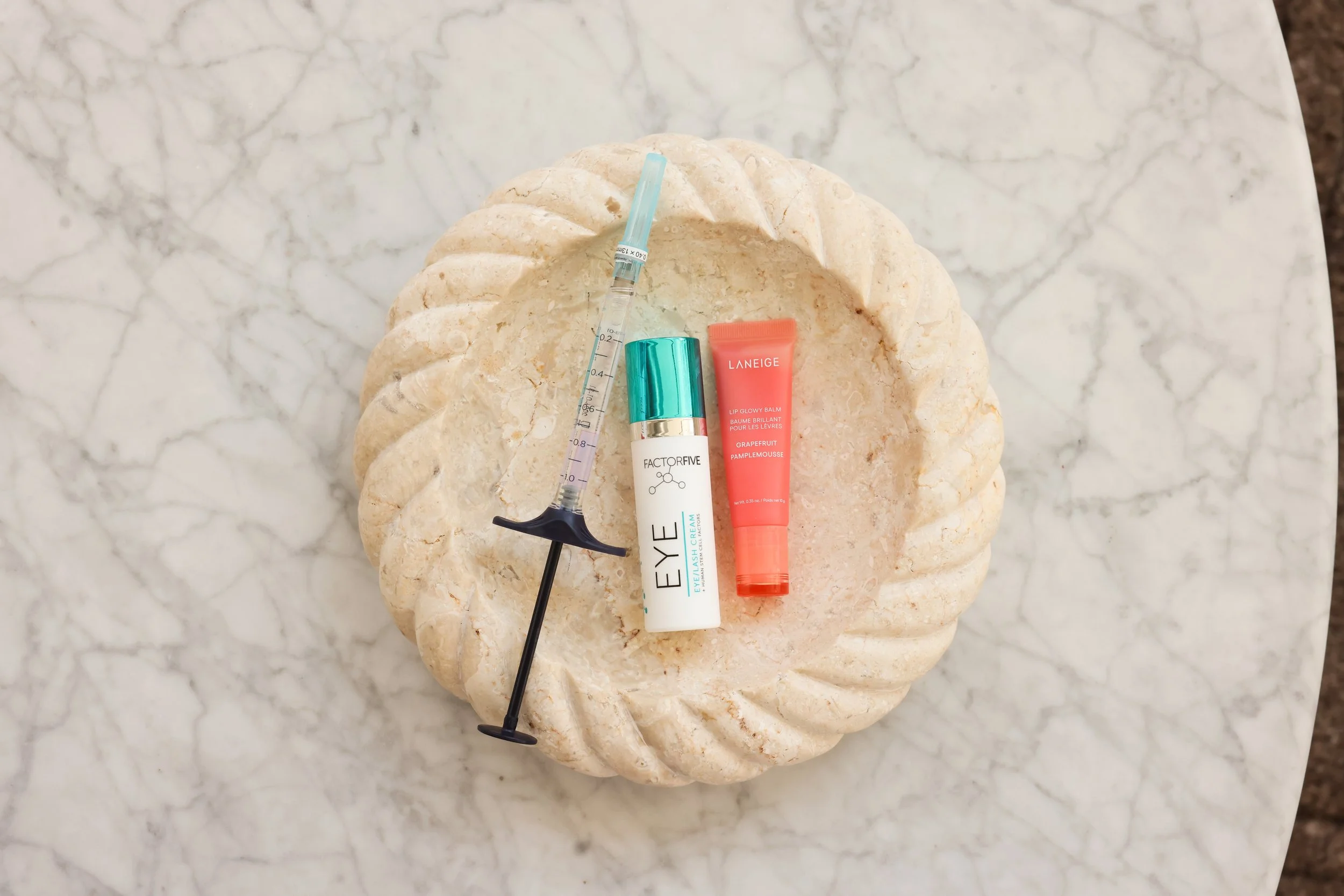 A decorative sculpture of a shell with three skincare products and a syringe placed on top of it. The background is a light-colored marble surface.