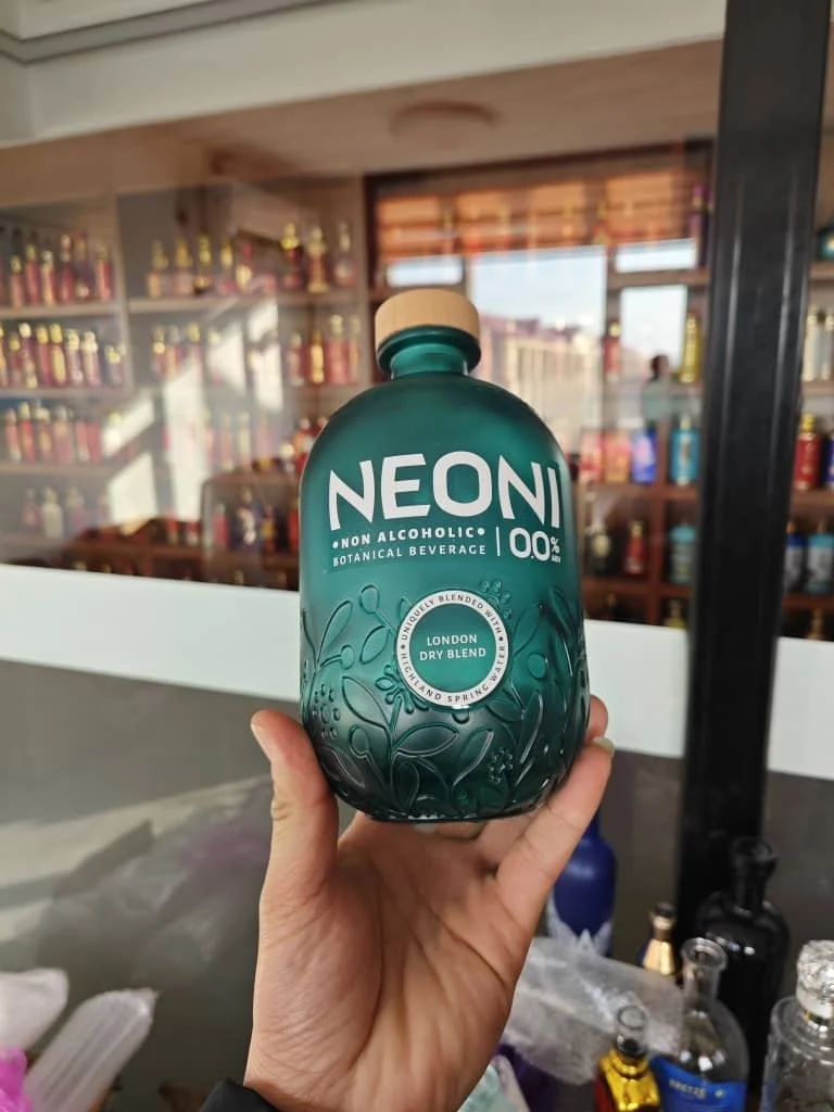A person holding a green bottle of NEON non-alcoholic botanical beverage with a wooden cap, label indicating London dry blend, in a store with shelves of various bottles in the background.