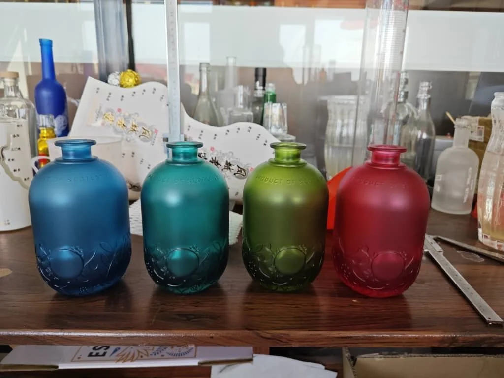 Four frosted glass perfume bottles in blue, teal, green, and red, lined up on a wooden surface in a laboratory or craft room.