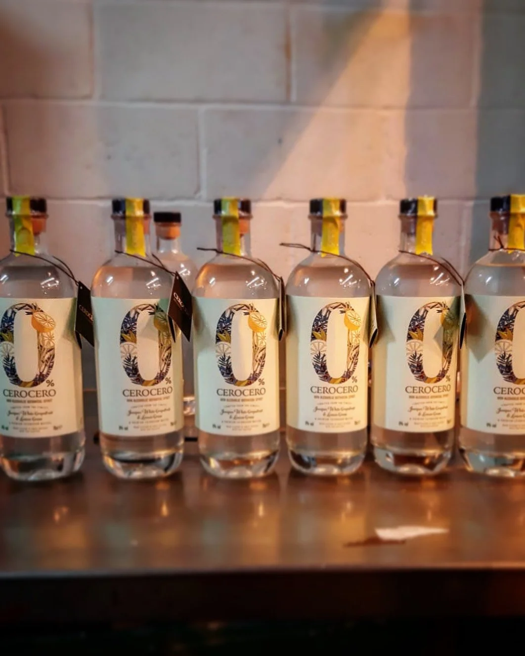 Six bottles of Cero Cero alcohol-free spirit lined up on a wooden surface with a brick wall background.