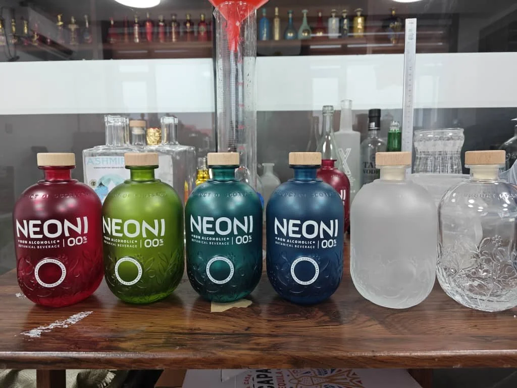 Colorful bottles of NEON non-alcoholic, botanical beverage on a wooden table, with a beaker and test tube behind them, and additional clear bottles to the right.