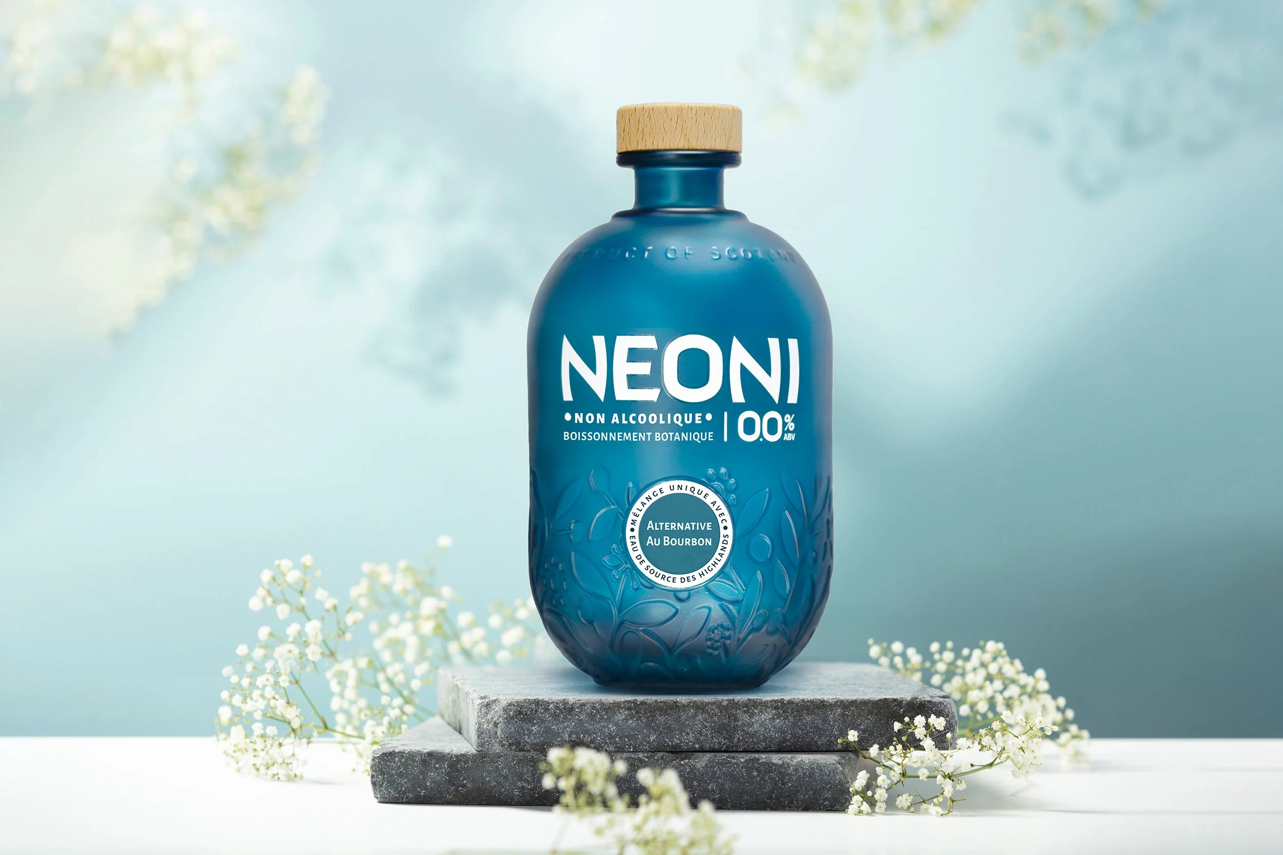 Blue bottled non-alcoholic beverage with wooden cap, standing on gray stone block surrounded by small white flowers, with a soft blue background.