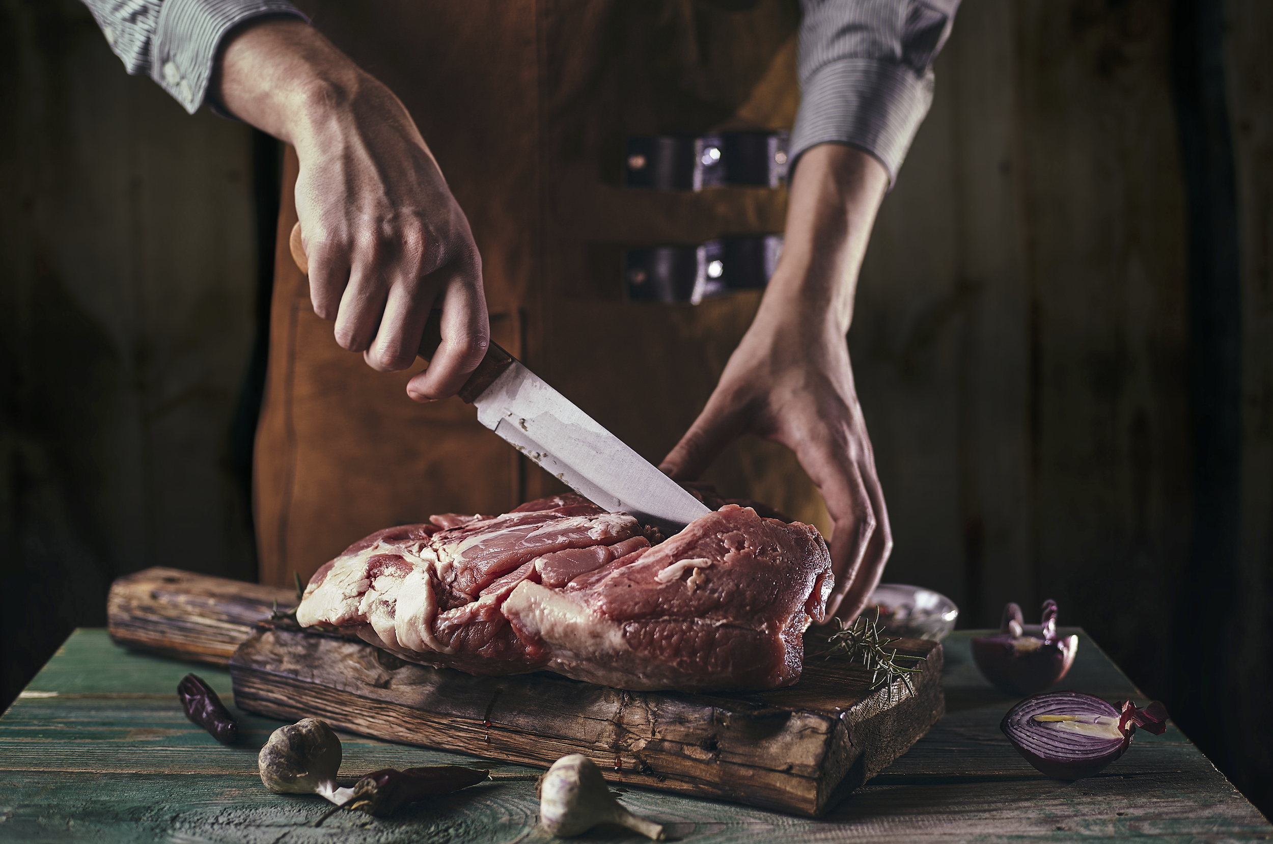 Person slicing raw meat on a wooden cutting board, surrounded by vegetables and herbs, with a rustic background.