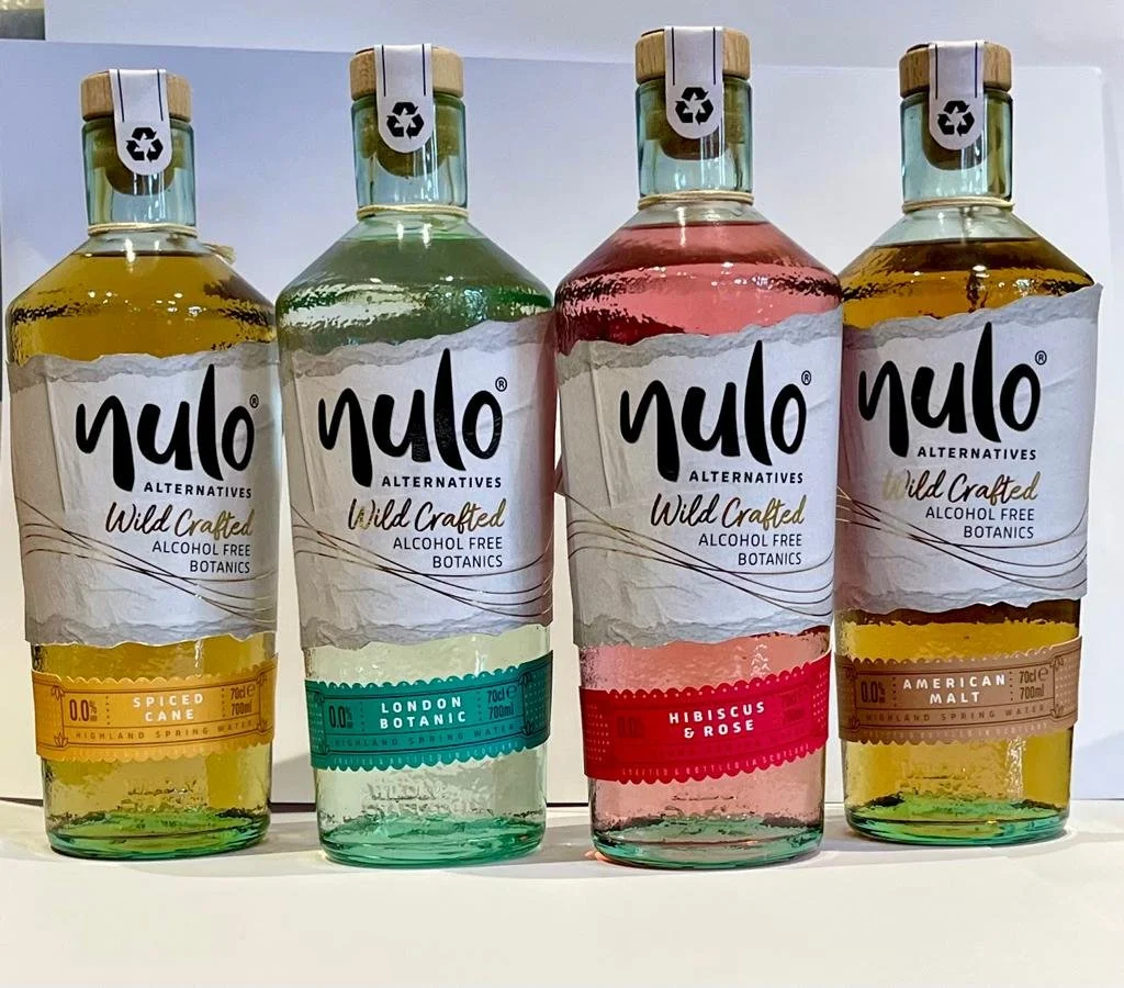 Four bottles of Nulo Alternatives alcohol-free botanic beverages in yellow, green, pink, and amber colors, labeled 'Spiced Cane,' 'London Botanic,' 'Hibiscus & Rose,' and 'American Malt' respectively, with wooden caps and white tags.