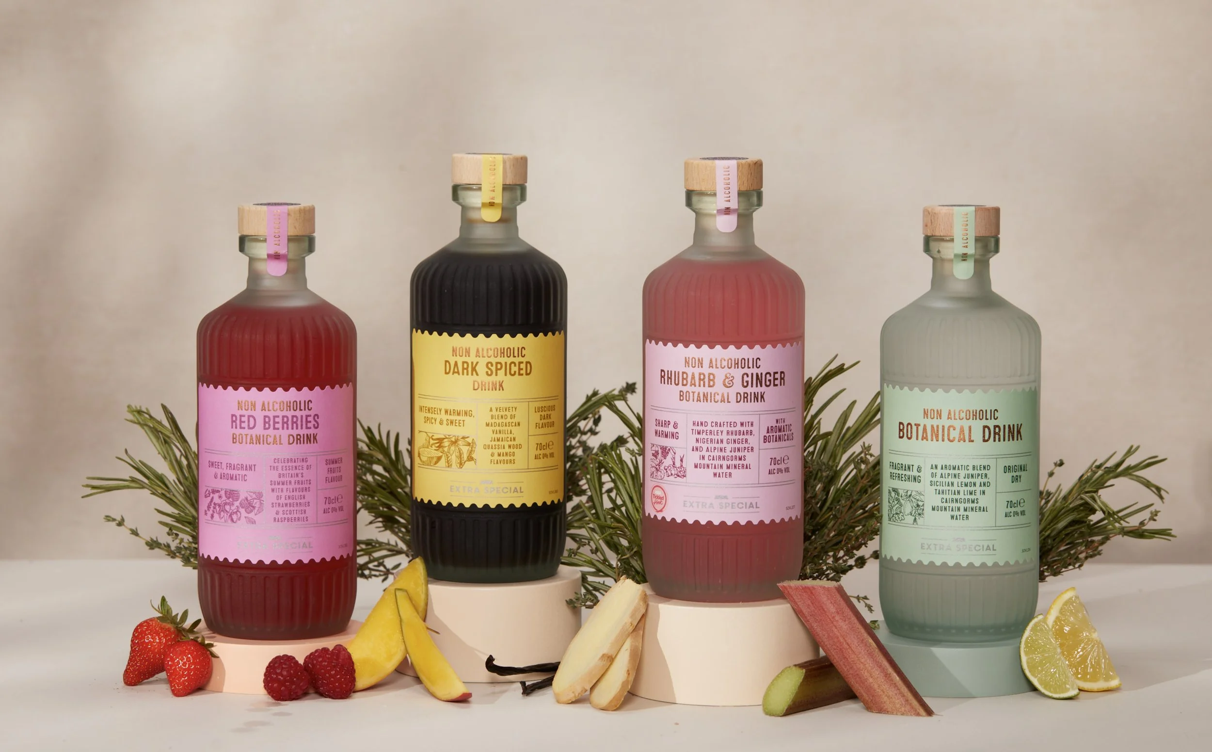 Four bottles of non-alcoholic botanical drinks with labels in pink, yellow, pink, and green, surrounded by fresh fruit slices and sprigs of greenery.