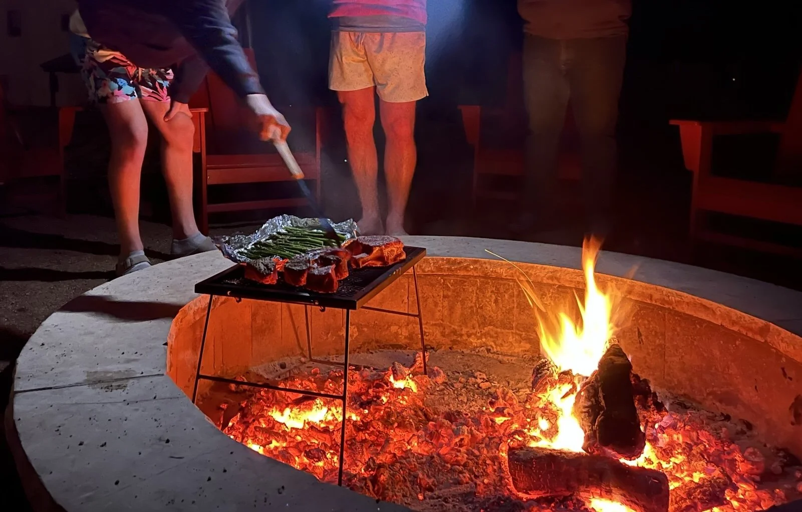 People cooking steaks and asparagus over an open fire in a fire pit at night.