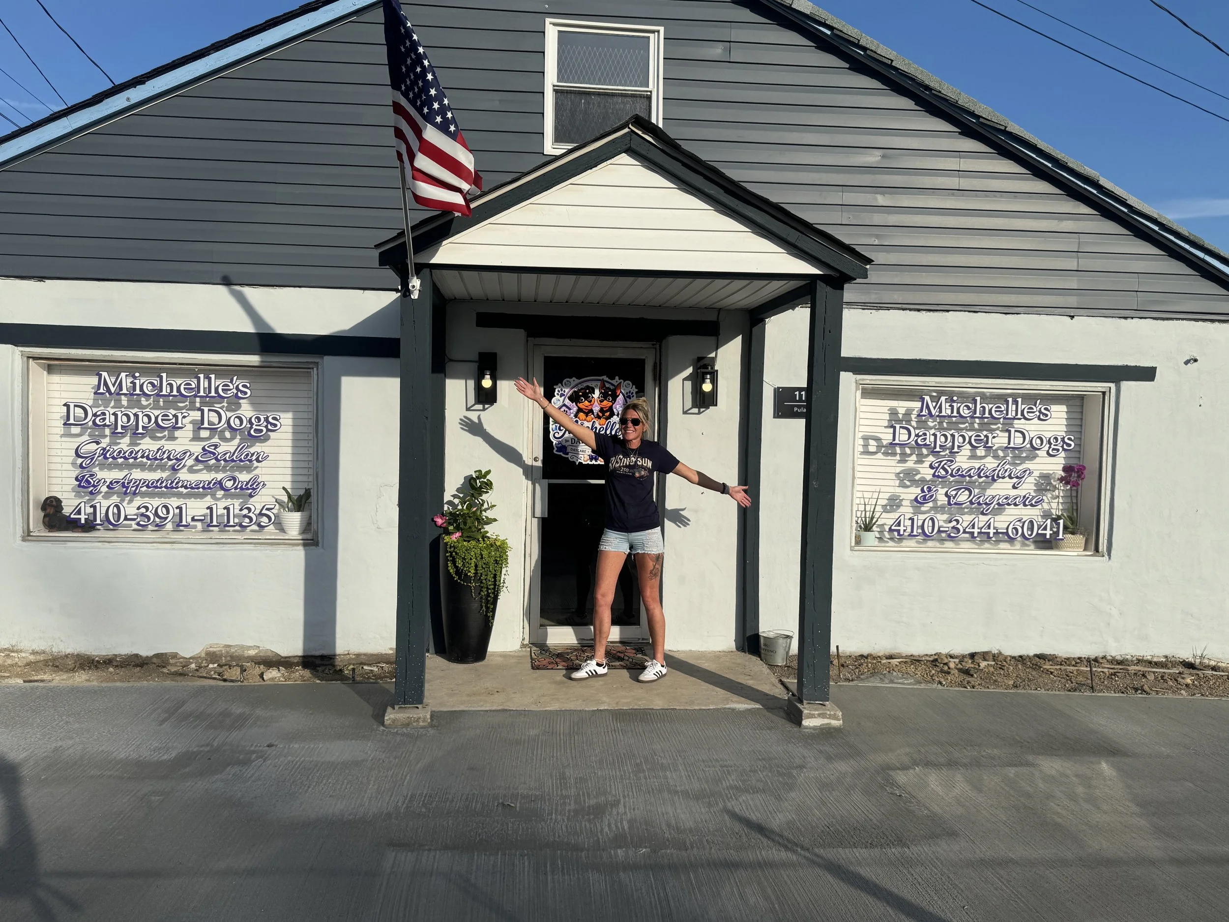 A woman standing in front of a grooming salon, waving and smiling, with an American flag flying above the building. The salon is called Michelle's Dapper Dogs, offering grooming, boarding, and daycare services.