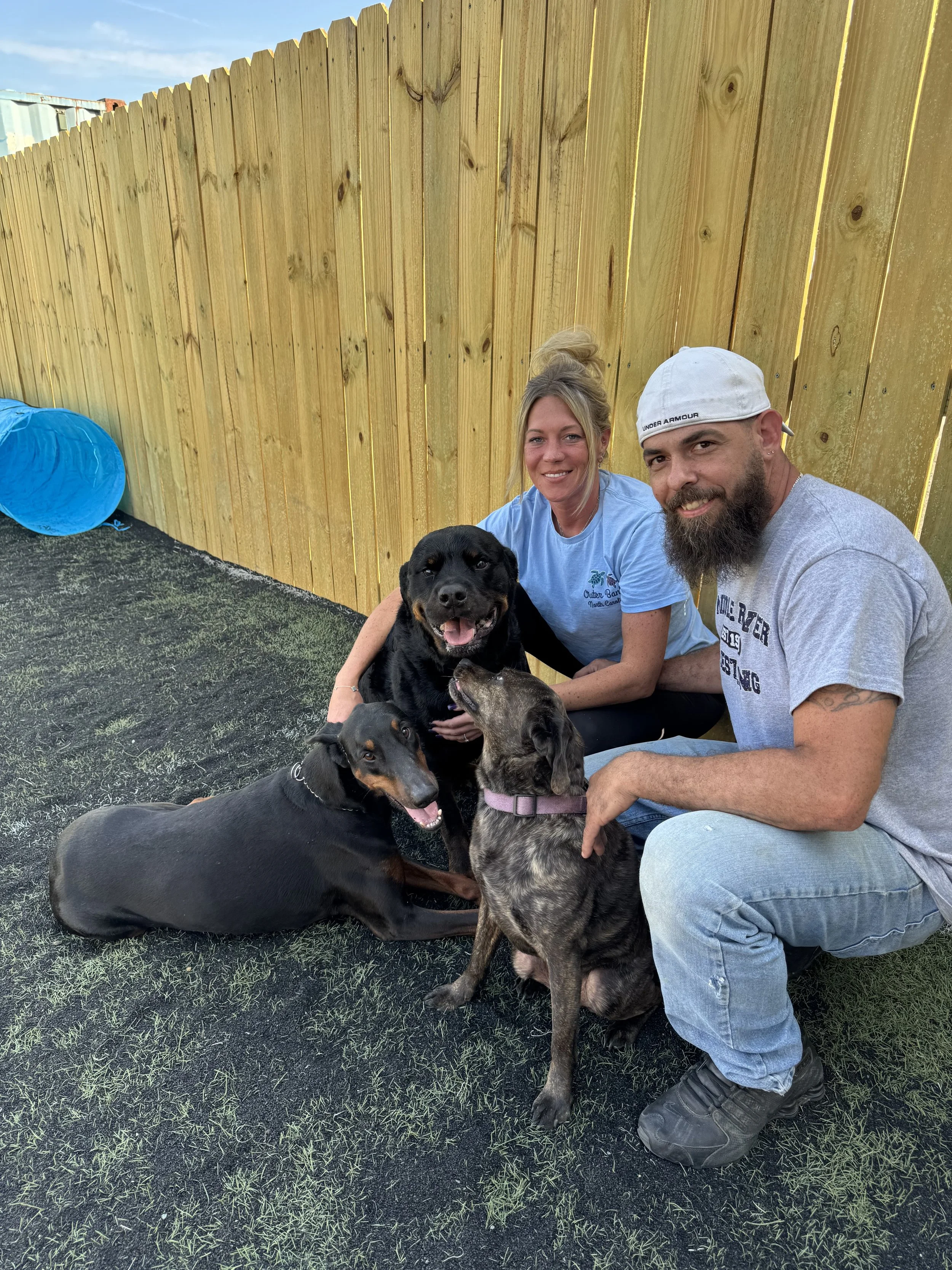 Michelle, owner, and Husband sitting on the ground in the front of a wooden fence with four dogs, including a black and tan dog, a brindle dog, and two black dogs, outdoors on a patch of grass and black top with a blue tunnel toy nearby.