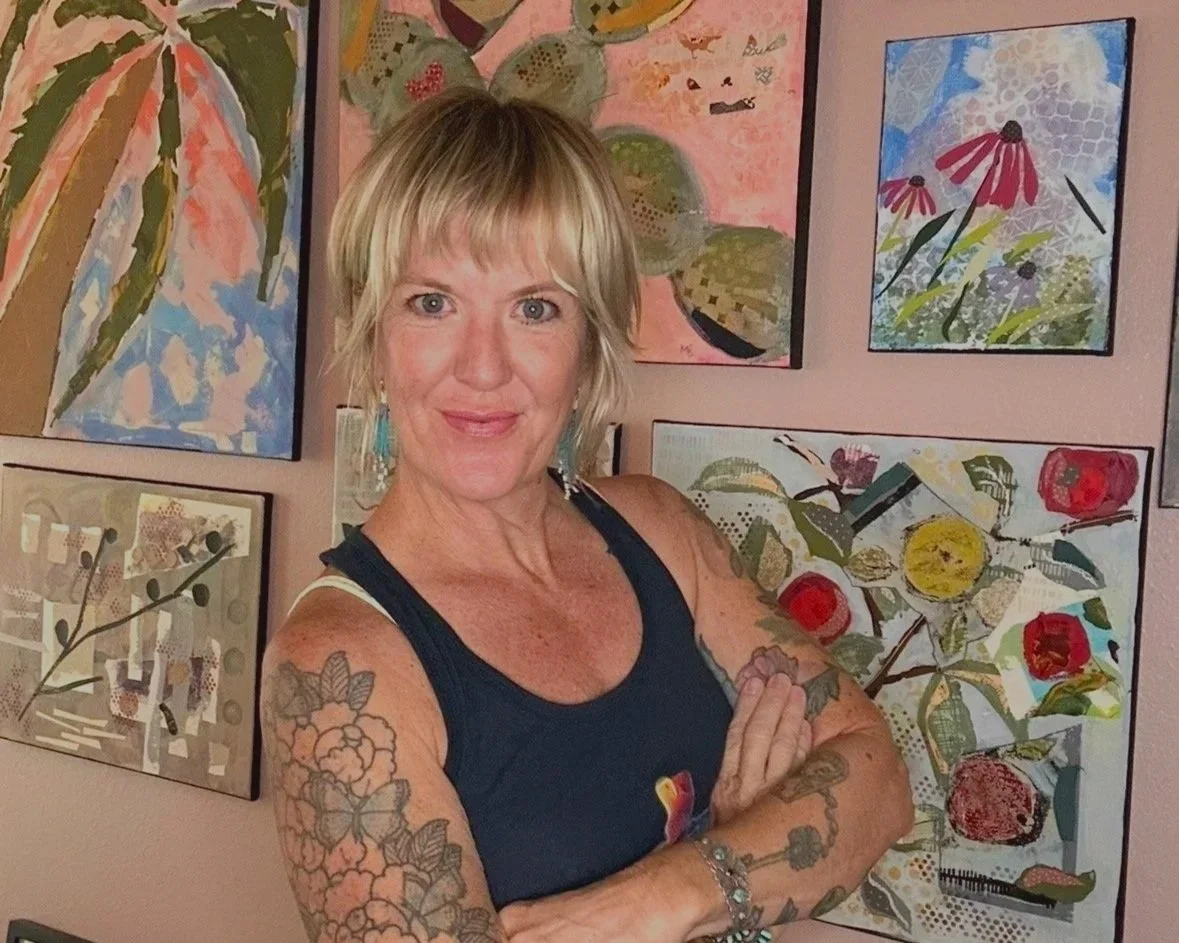 Woman with tattoos on her arms and earrings, standing with arms crossed in front of a colorful wall of abstract floral paintings.