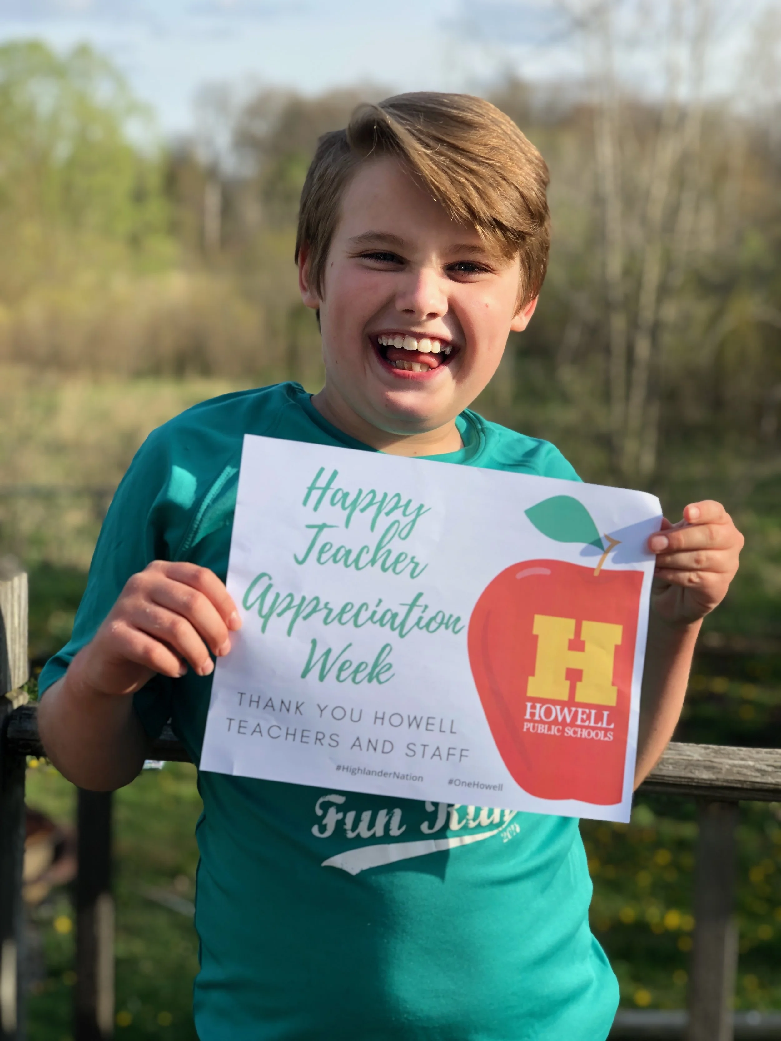 Smiling boy holding a sign that says 'Happy Teacher Appreciation Week' with the Howell Public Schools logo in an outdoor setting.