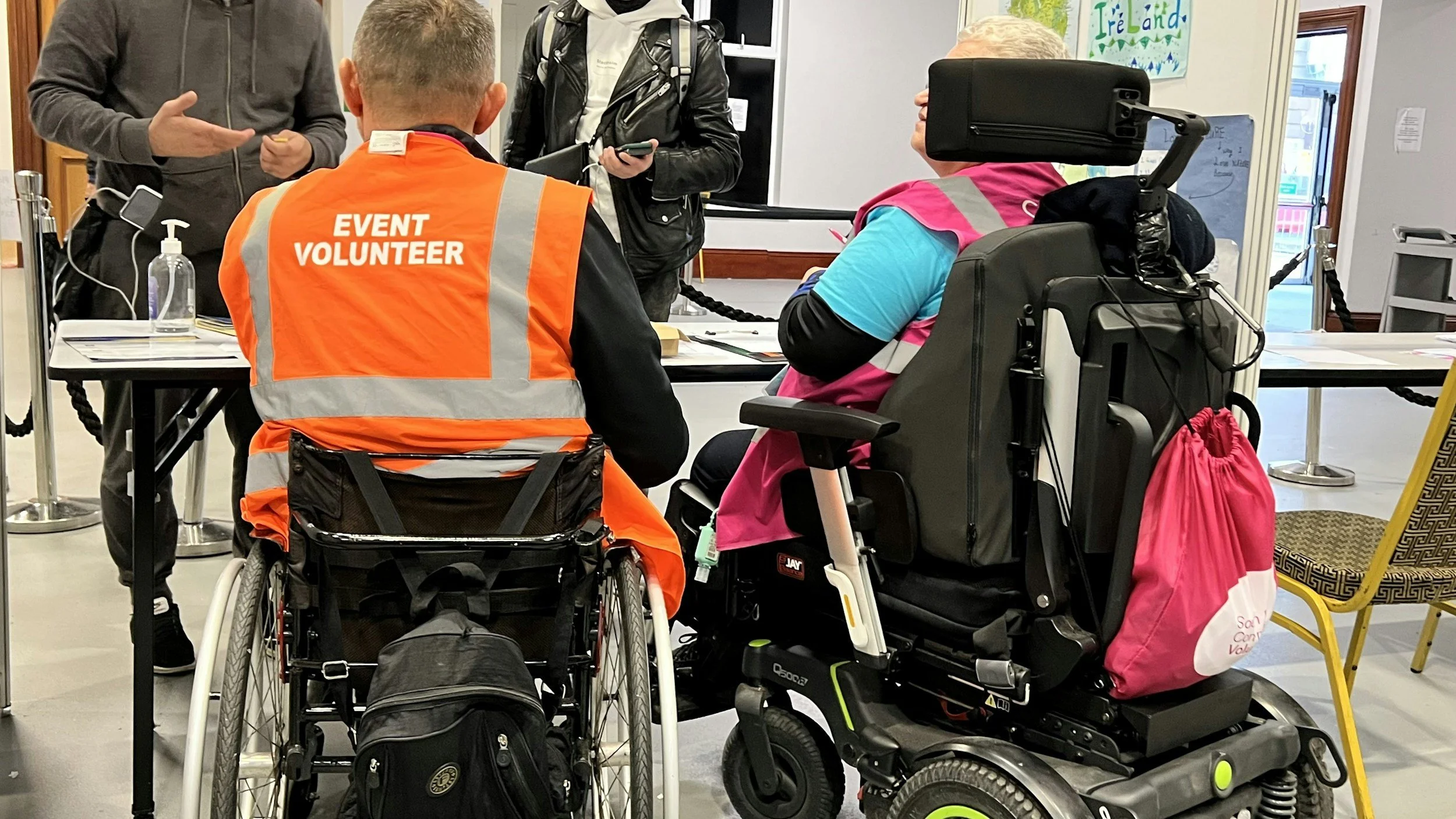 People at a check-in or registration desk, including a person in a wheelchair and another in a motorized wheelchair, wearing a pink and blue jacket, with an event volunteer in an orange vest, and others standing nearby.