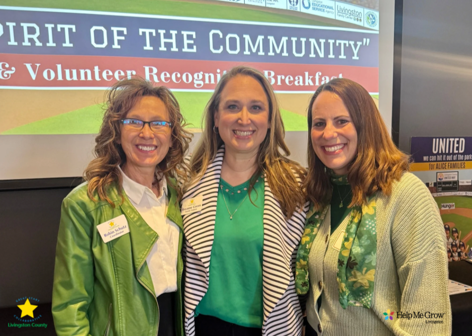Three women standing together at an event with a screen behind them. The screen displays the words "Spirit of the Community" and mentions a volunteer recognition breakfast. The women are smiling, wearing name tags and green clothing.