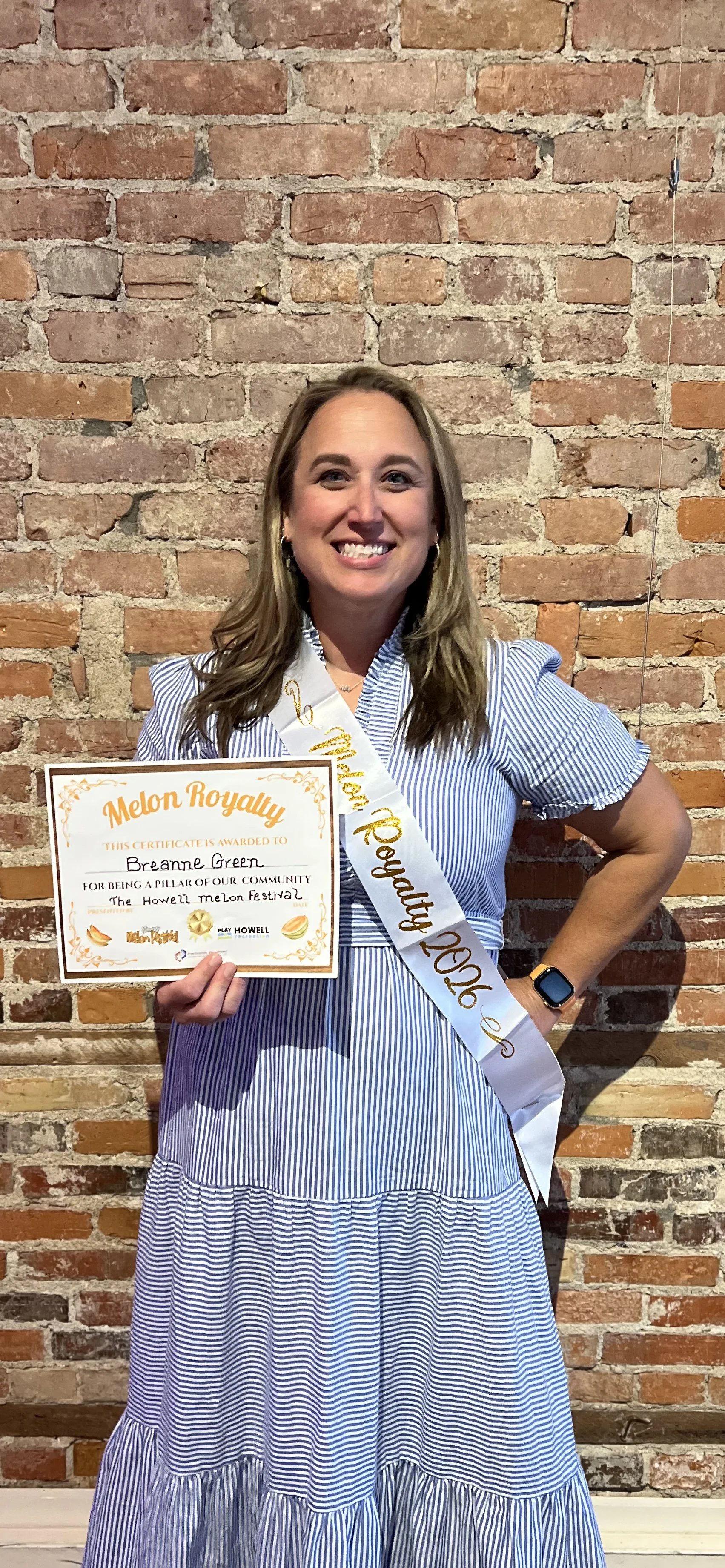 Woman in blue and white striped dress, wearing a satin sash that says 'Melon Royalty 2023,' smiling, holding a certificate, standing in front of a brick wall.
