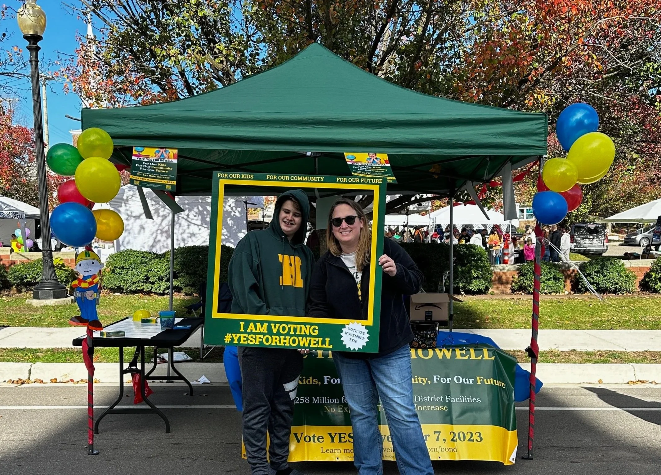 Two people standing under a green tent at an outdoor voting booth, holding a large green and yellow sign encouraging voting for Howell, with colorful balloons decorating the tent, and a crowd in the background.