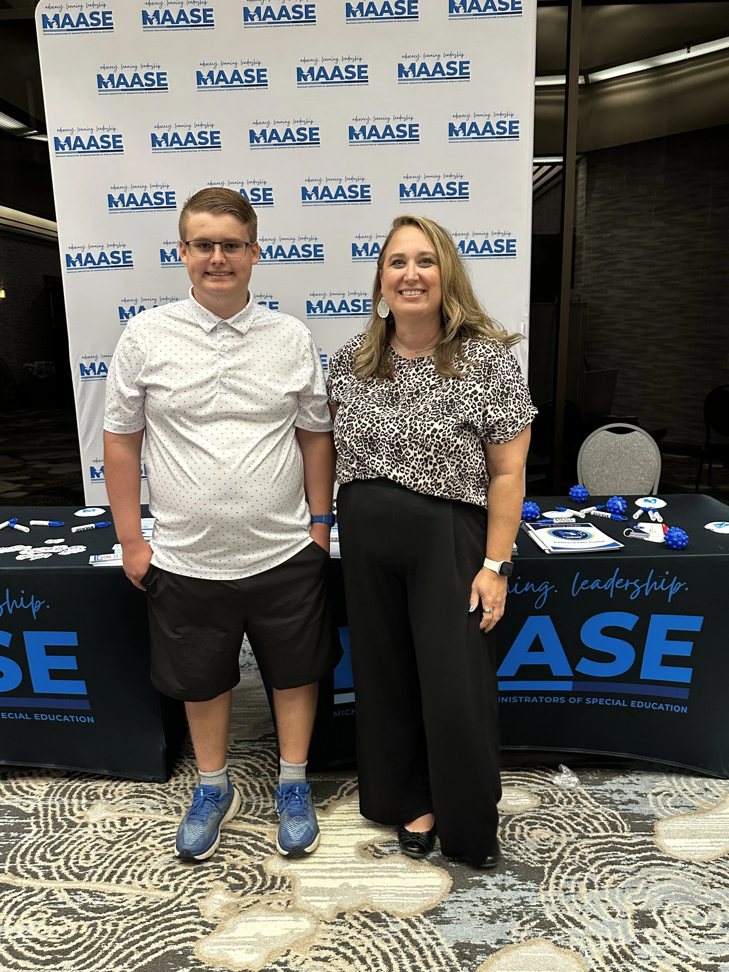 A young man and a woman standing side by side at a conference, in front of a backdrop with the logo and text of the Michigan Association of Administrators of Special Education (MAASE). The woman is smiling, wearing a leopard print blouse and black skirt, and the young man is wearing glasses, a white polka dot shirt, and black shorts.
