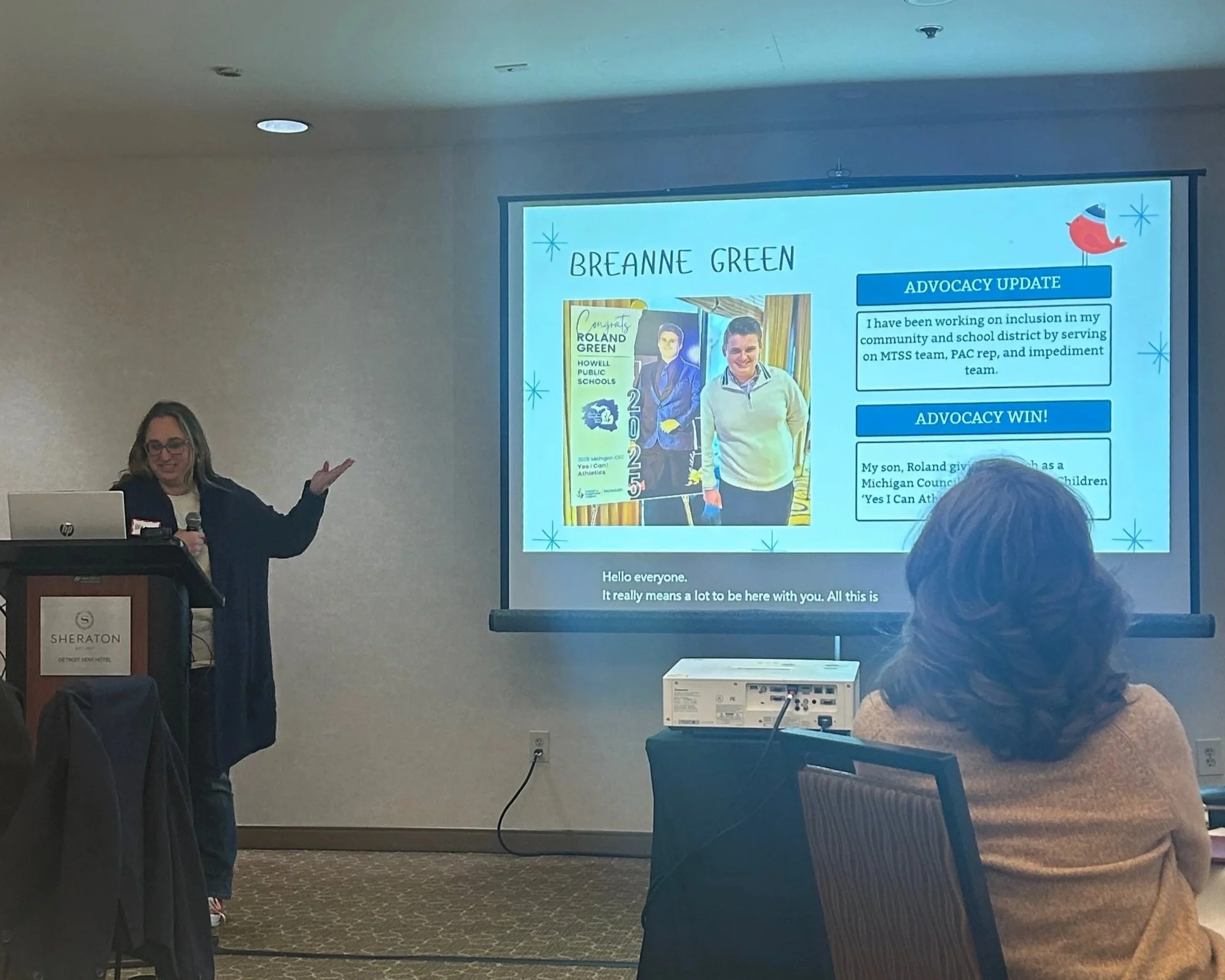 A woman presenting in front of a large screen at a conference or meeting room. The screen displays a slide with the title "BREANNE GREEN" and an advocacy update. The slide includes an image of a young person and some text. Part of the audience is visible, with one person in the foreground listening.