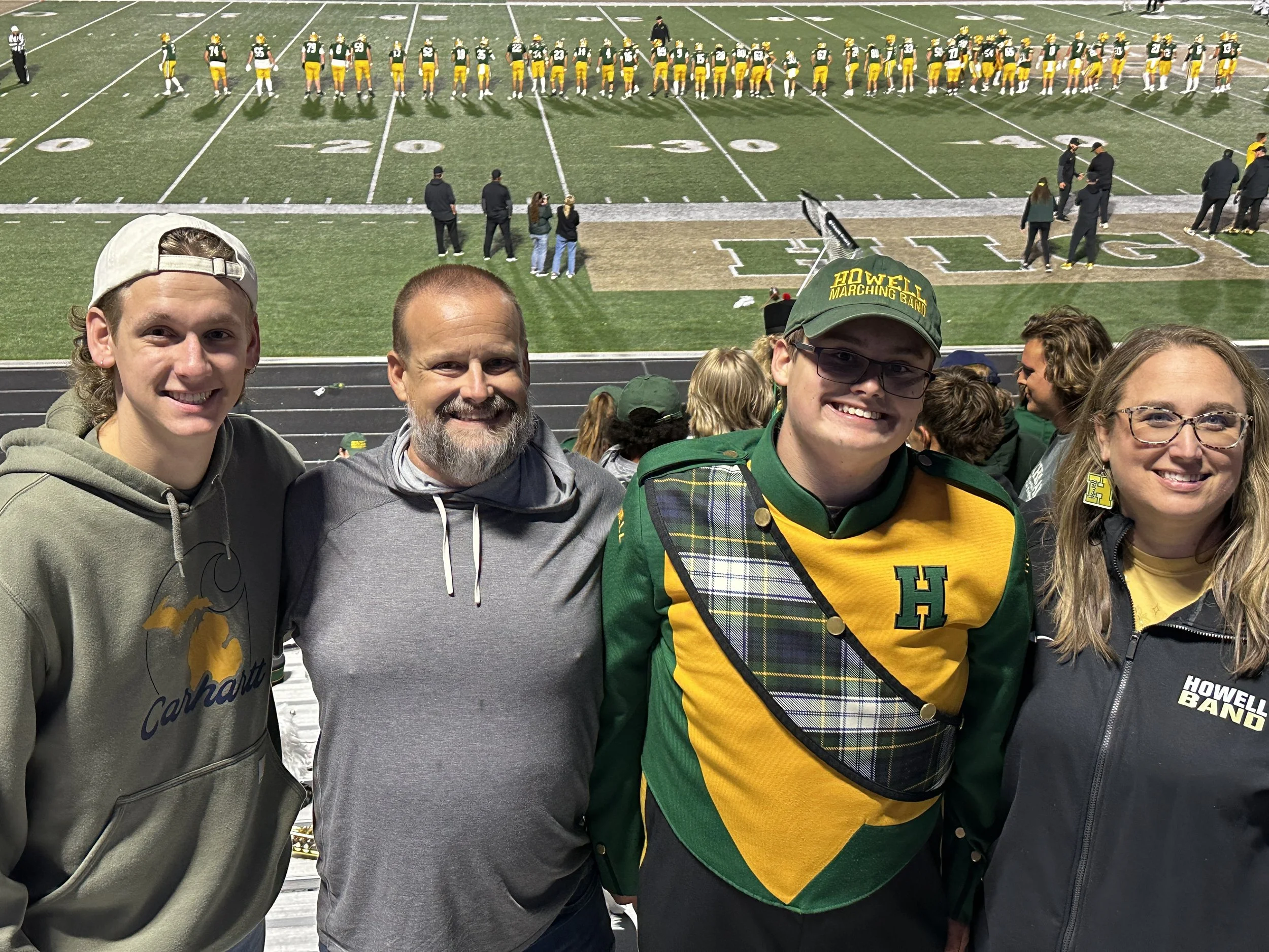 Four people standing together at a football game, with a football field and players in the background. The person on the far right is wearing a hoodie with the Carhartt logo, the second person from the left has a beard and gray hoodie, the third person from the left is wearing glasses and a green and yellow outfit representing the Howells Band, and the woman on the far right is wearing glasses and a jacket with a band patch.