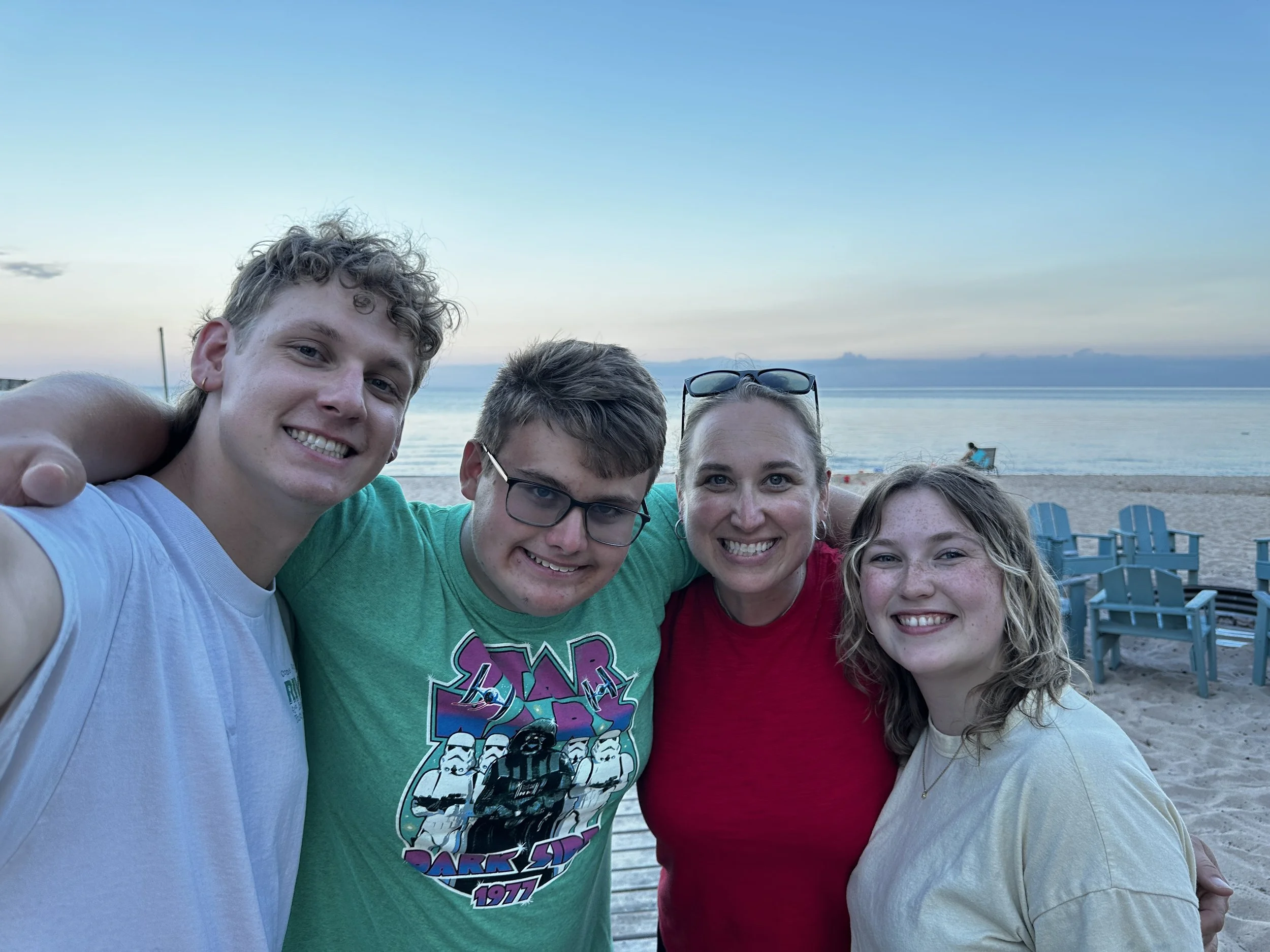 A group of five friends smiling and taking a selfie on the beach with an ocean view and blue chairs in the background.