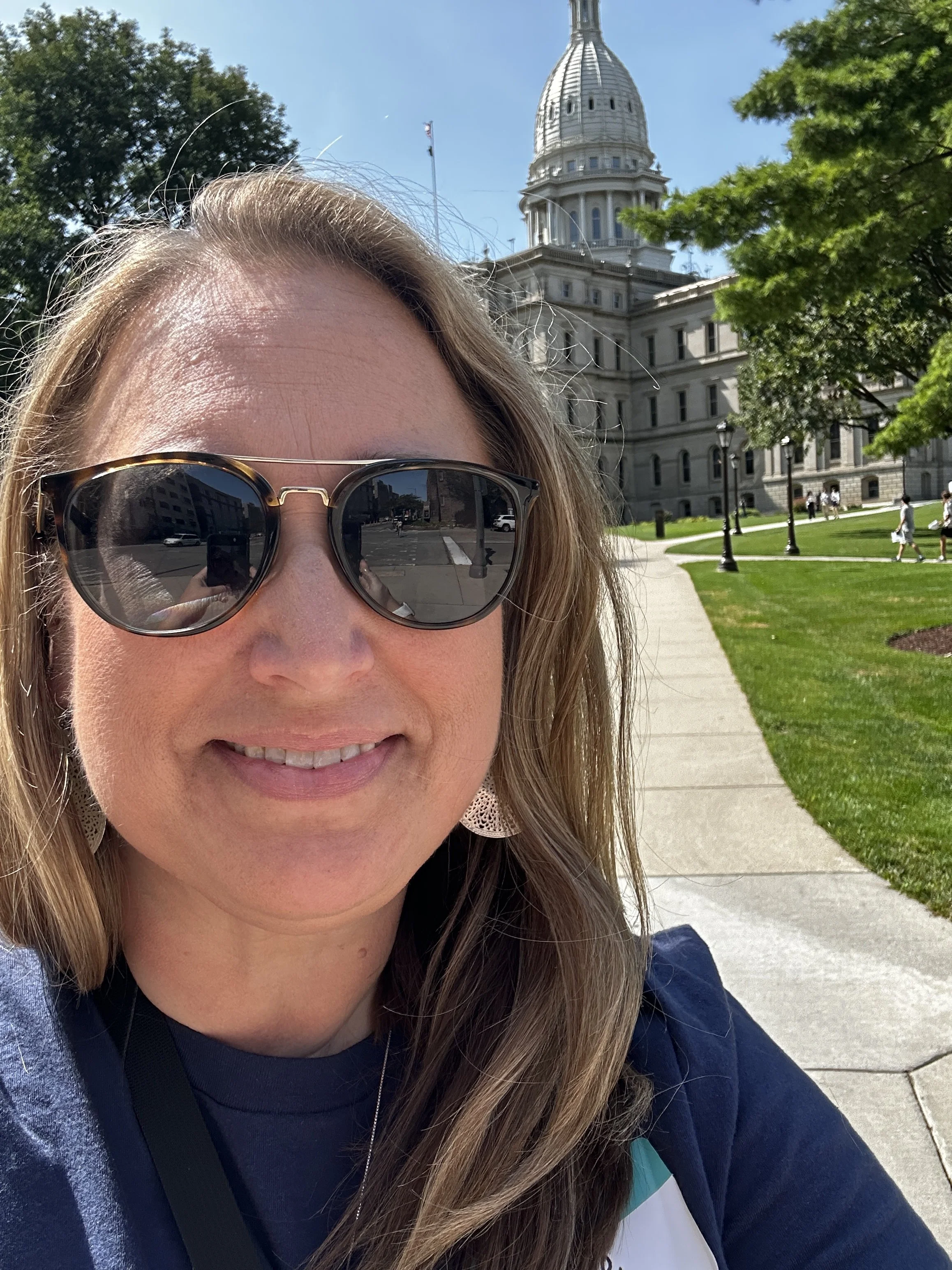 Close-up of a smiling woman wearing sunglasses with a government building with a dome in the background, outside in a sunny park with trees and a sidewalk.