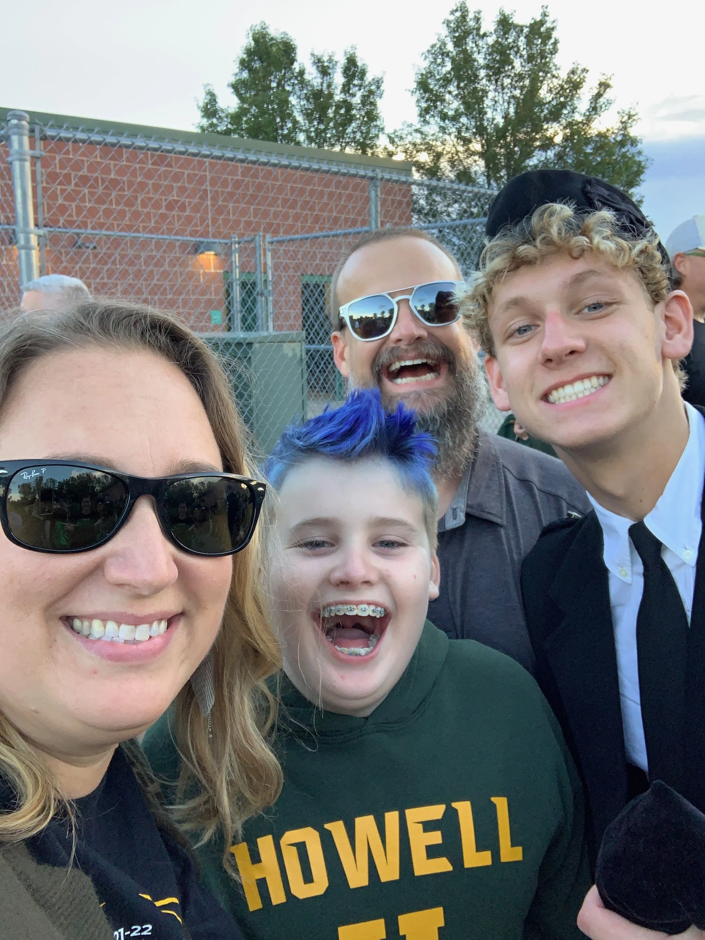 Group of smiling people taking a selfie outdoors near a chain-link fence and trees at sunset.