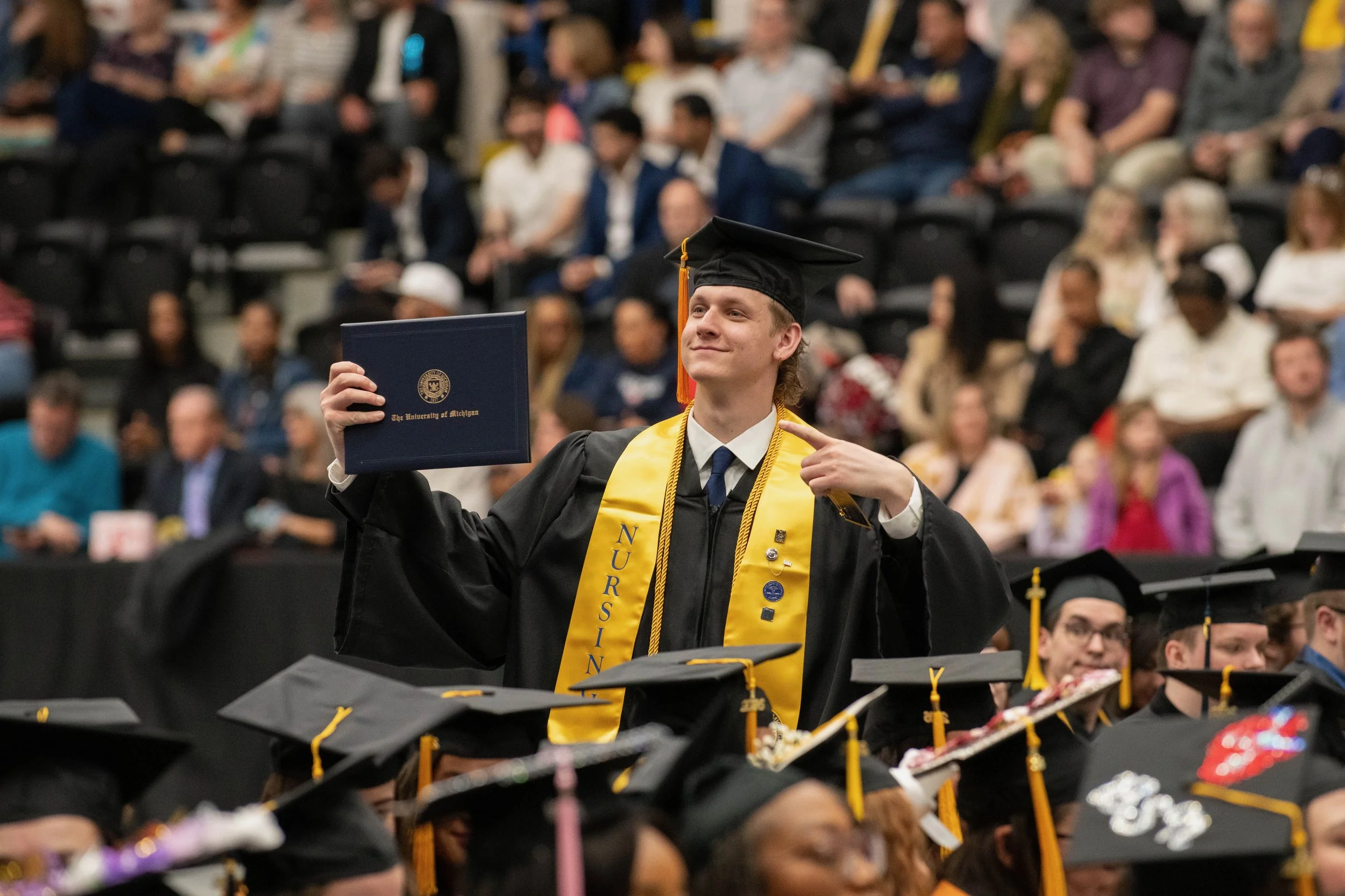 A young man in graduation attire, including a black cap and gown with a yellow stole, holding a diploma in one hand and pointing at himself with the other, during a ceremony attended by many people in a large indoor venue.