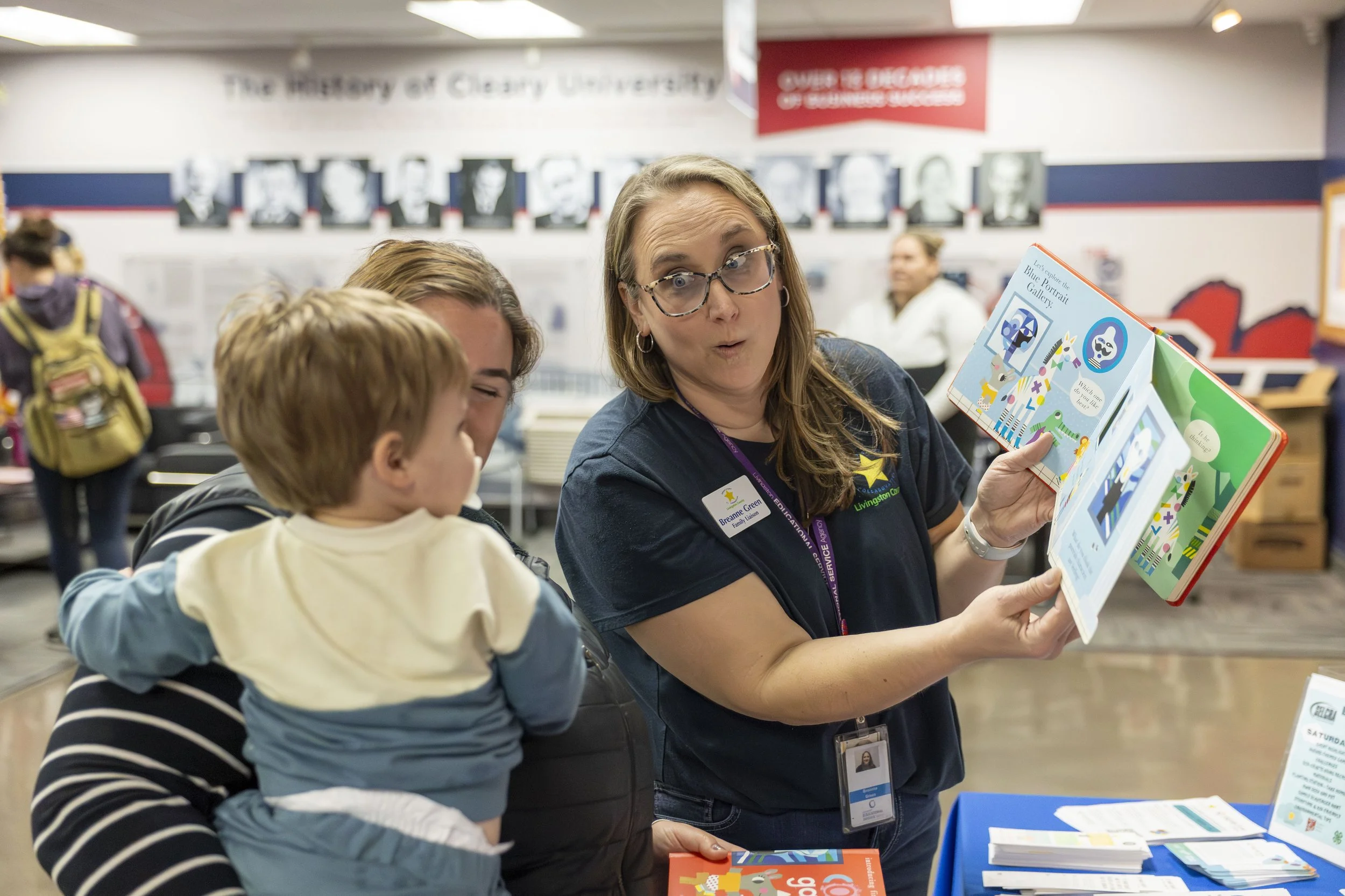 A woman showing a children’s book about the Blue Portrait Gallery to a young child at a library or bookstore. The woman is wearing glasses and a badge, and there are other people in the background.