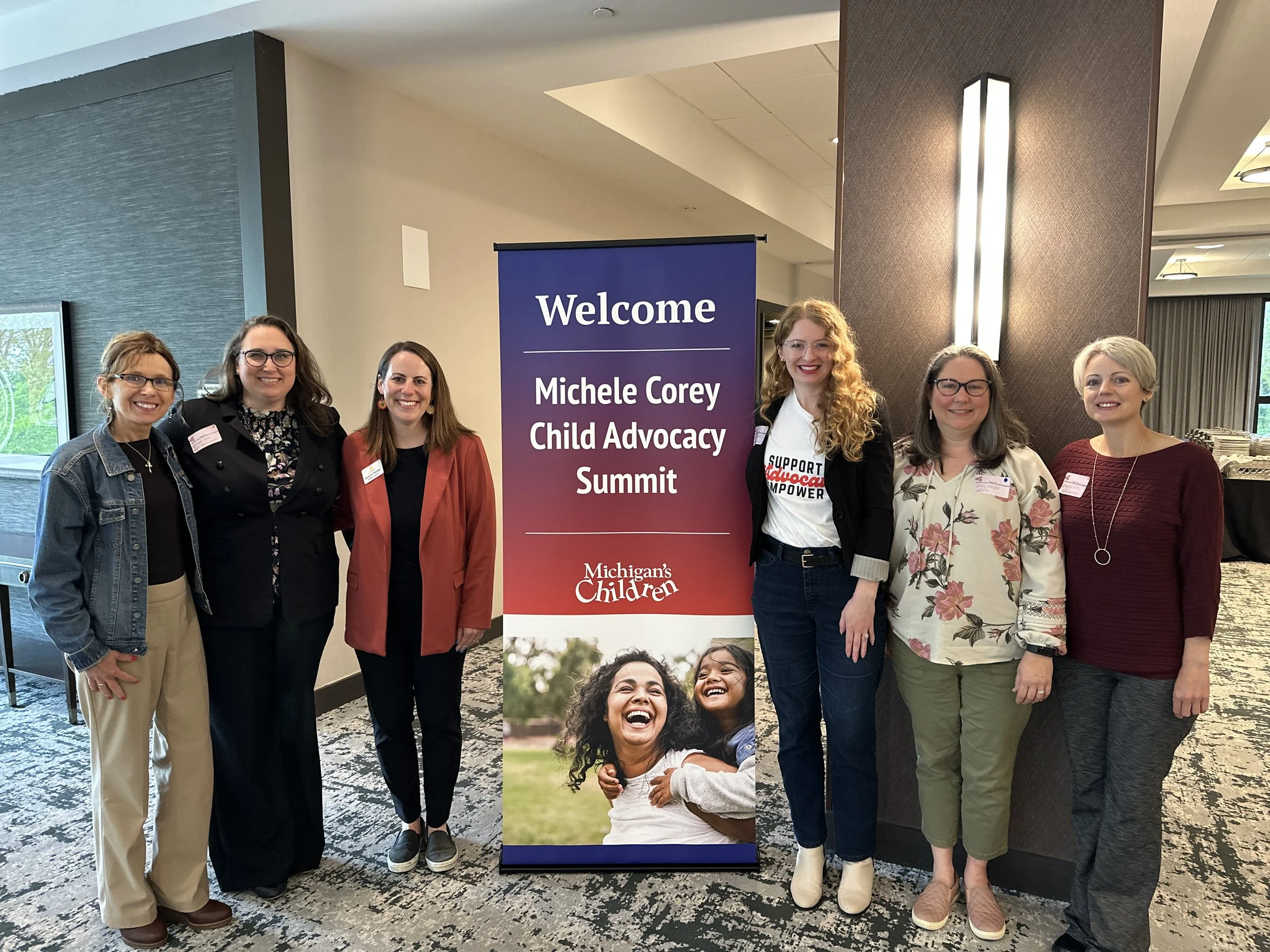 Group of seven women standing in front of a banner that reads "Welcome Michele Corey Child Advocacy Summit" at a conference or event. They are smiling and dressed in casual to business casual attire.