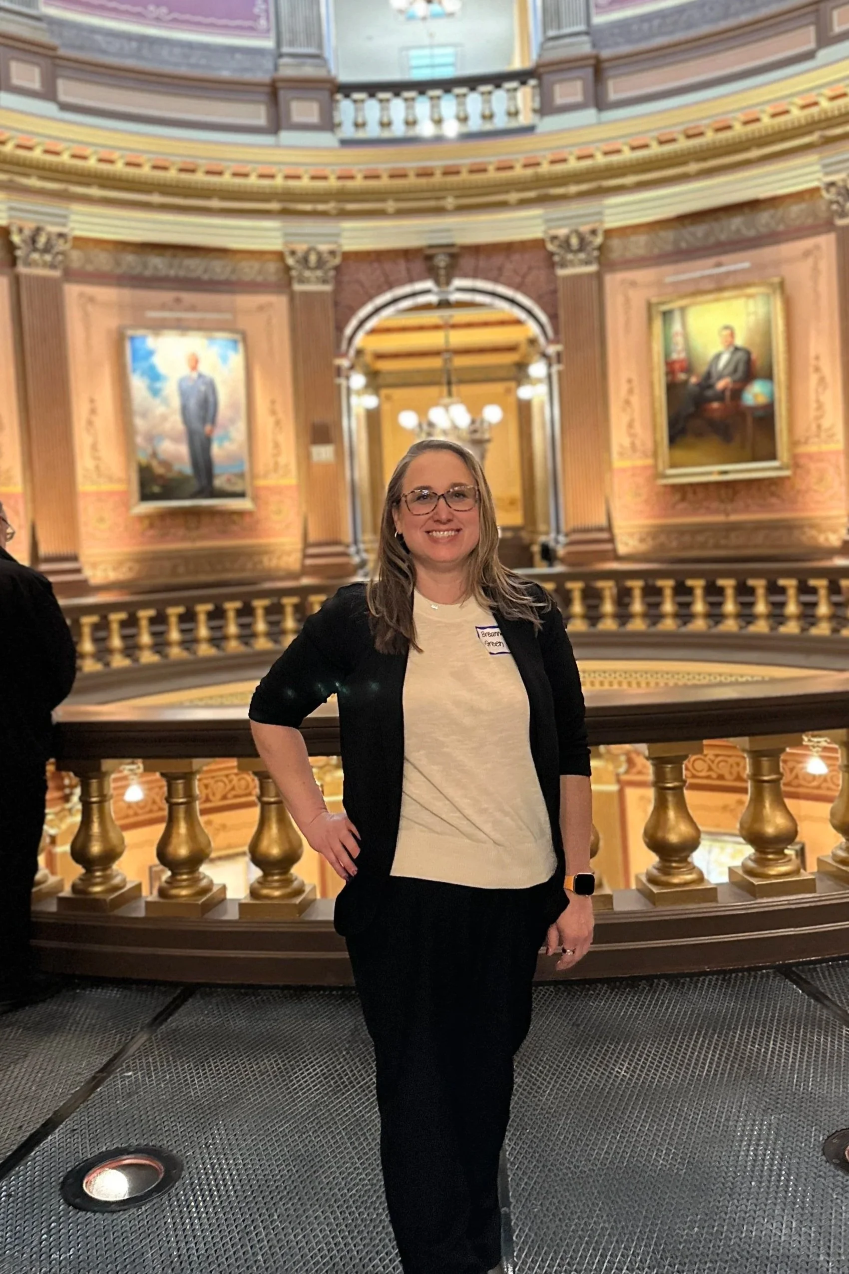 A woman with glasses and long hair wearing a beige top and black cardigan standing in front of a grand, ornate interior with portraits and gold railings.